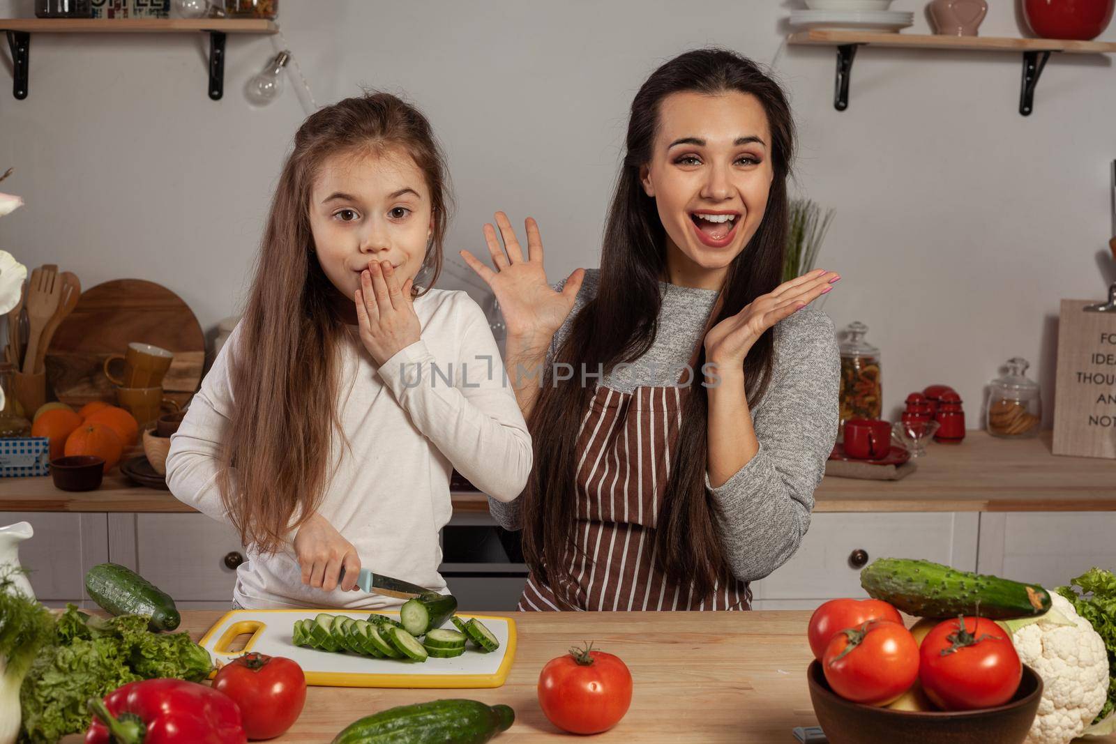 Happy loving family are cooking together. Cute mommy and her little girl are making a vegetable salad and having fun at the kitchen, against a white wall with shelves and bulbs on it. Homemade food and little helper.