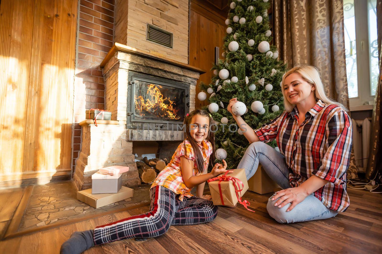 Mother holds a little daughter at the Christmas tree on a festive evening in a rustic wooden house, New Year, winter time