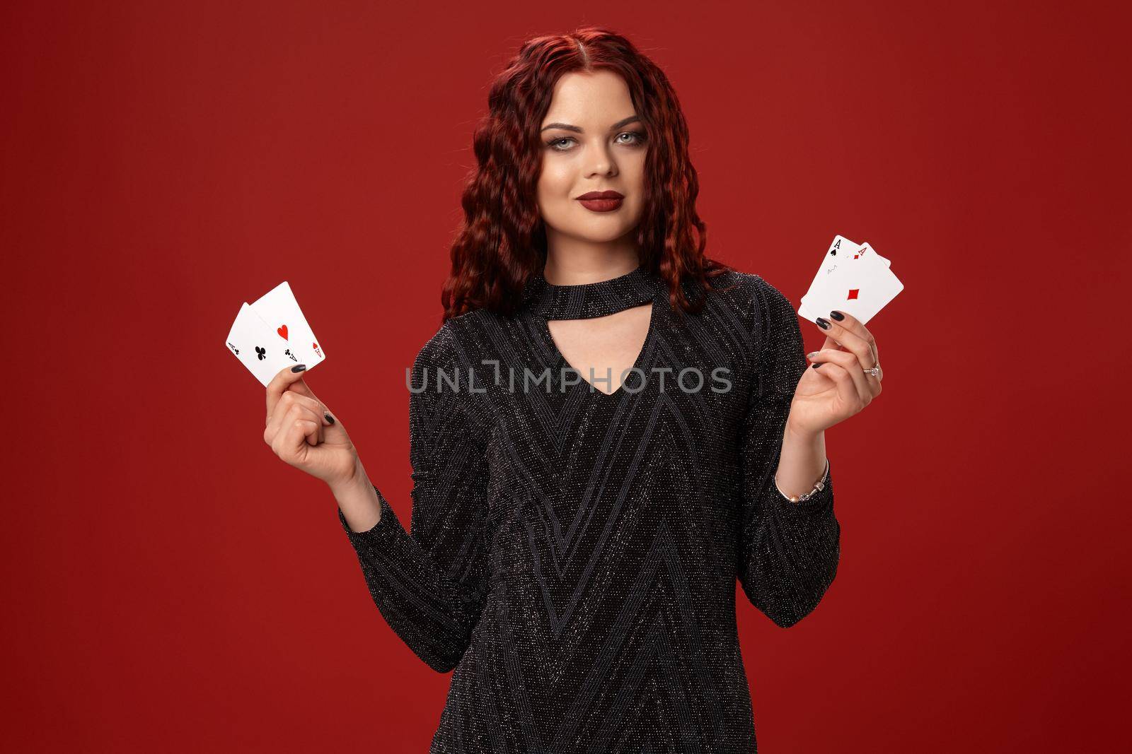 Beautiful redheaded woman in a black knitted dress is holding some playing cards and smiling. Casino. Four aces. Studio shot on a red background.