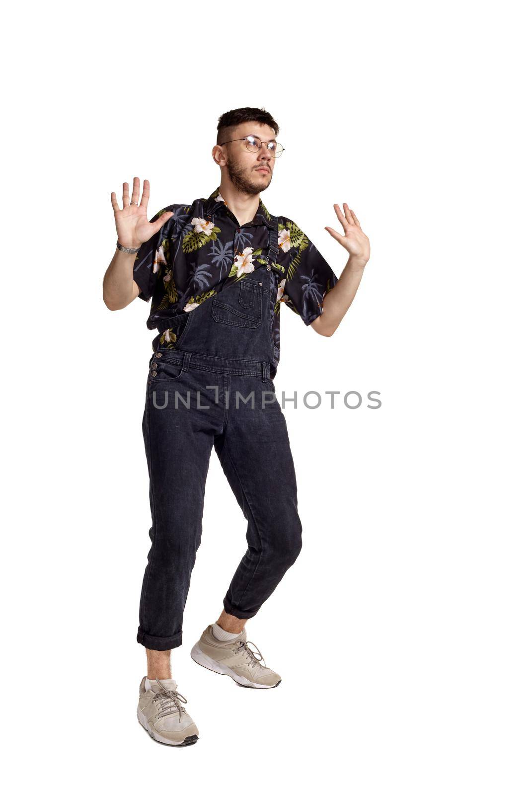 Full-length portrait of a good-looking person in glasses, black jumpsuit, colorful t-shirt and gray sneakers fooling around in studio. Indoor photo of a man dancing isolated on white background. Music and imagination concept.