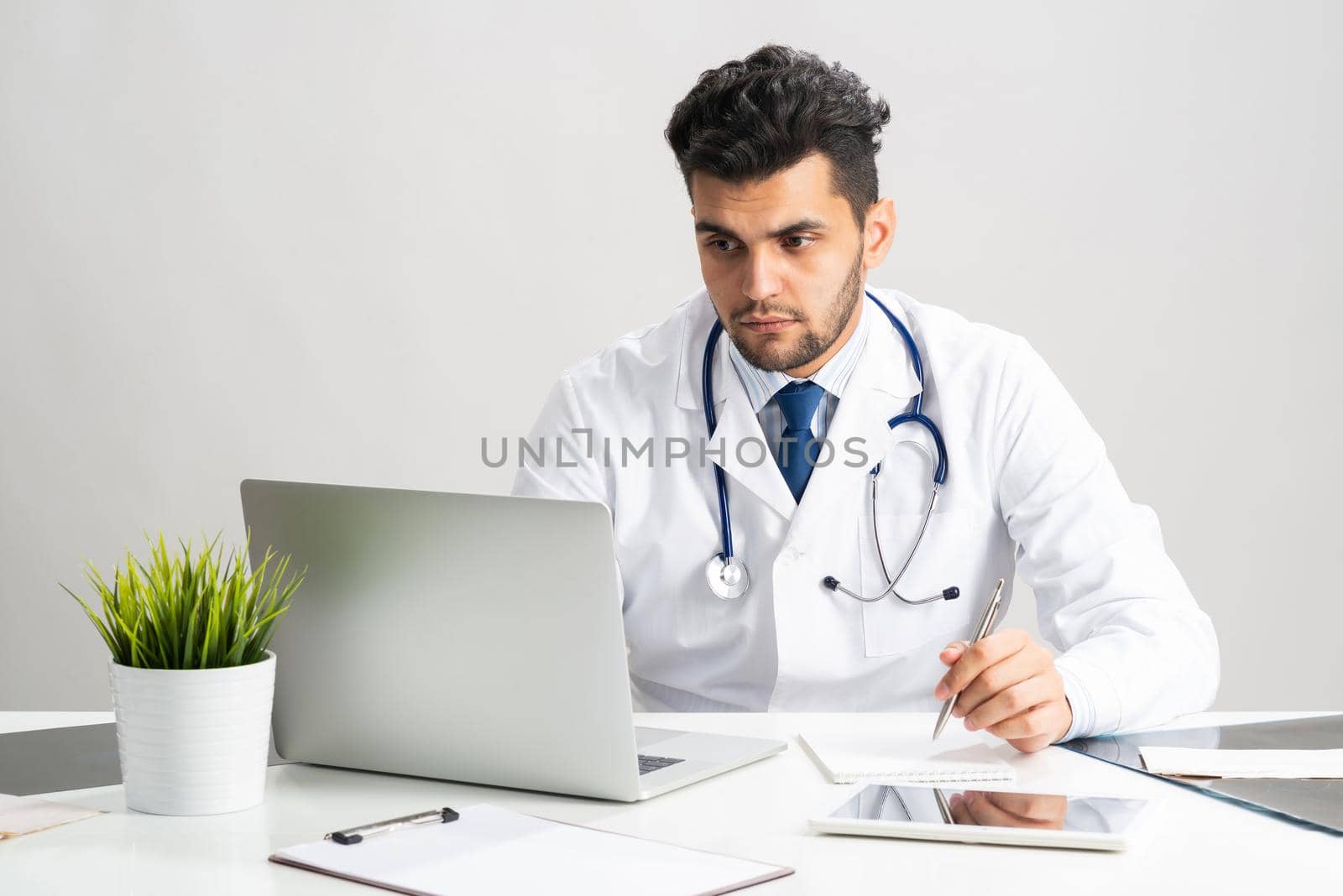 Young arab internist working at computer. Physician in white coat with stethoscope sitting at desk with laptop. Professional medical consultation and treatment in modern clinic. Medical healthcare