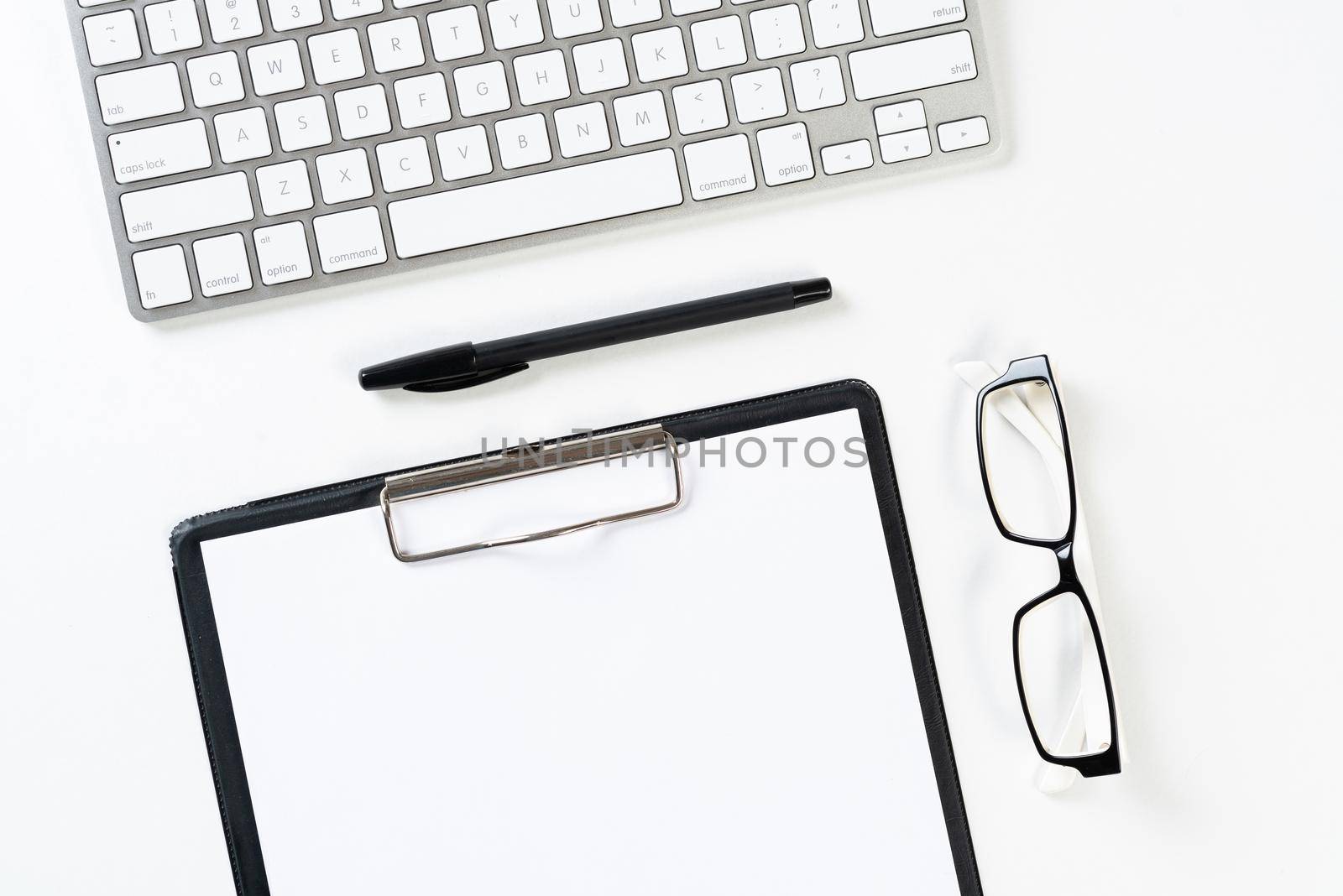 Top view of well organized workplace with office accessories. Clipboard with blank paper page, glasses and wireless keyboard on white surface. Education, creativity and working concept with copy space