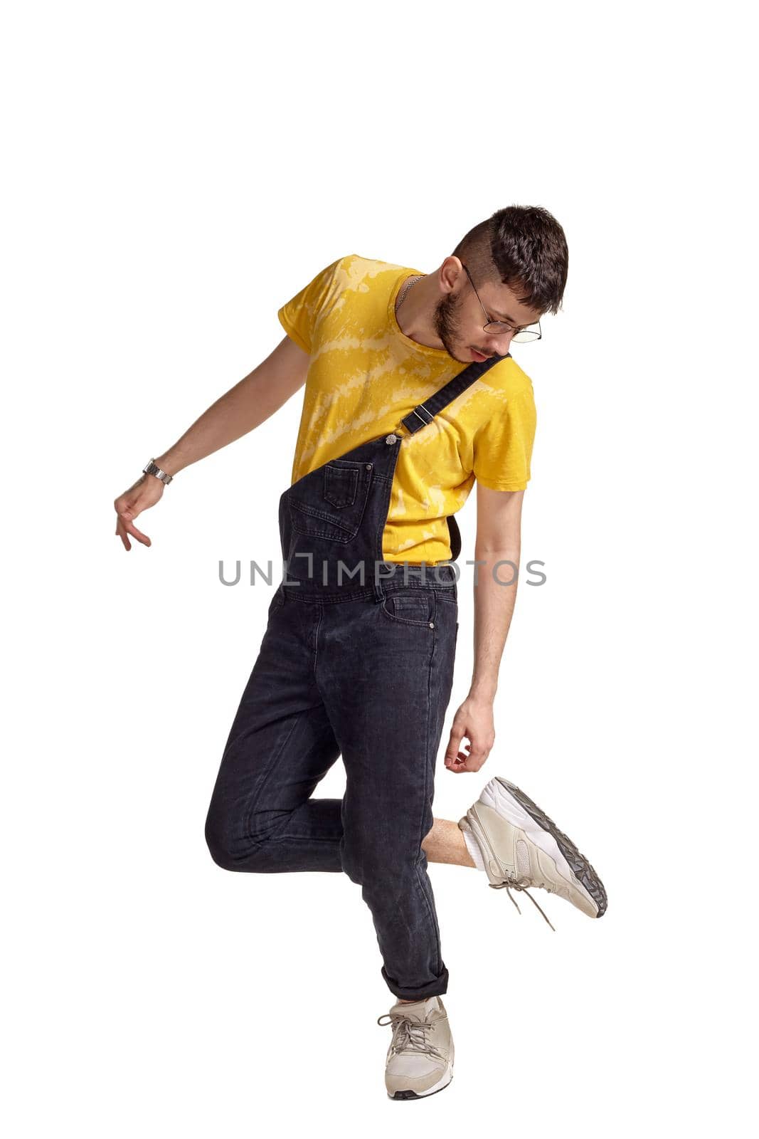 Full-length portrait of an athletic man in glasses, black jumpsuit, yellow t-shirt and gray sneakers fooling around in studio. Indoor photo of a man making dancing elements isolated on white background. Music and imagination concept.