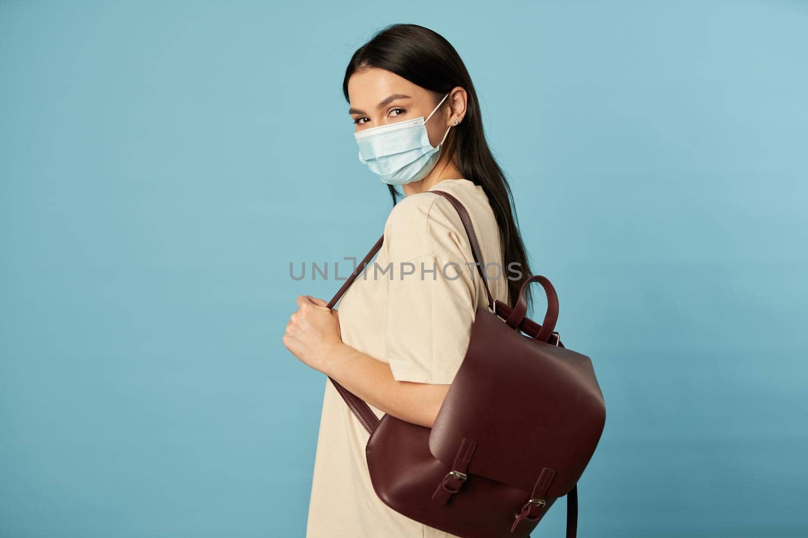 Side view of young lady in face mask holding backpack and looking at camera in studio, isolated on blue background. Copy space. Quarantine, epidemic concept