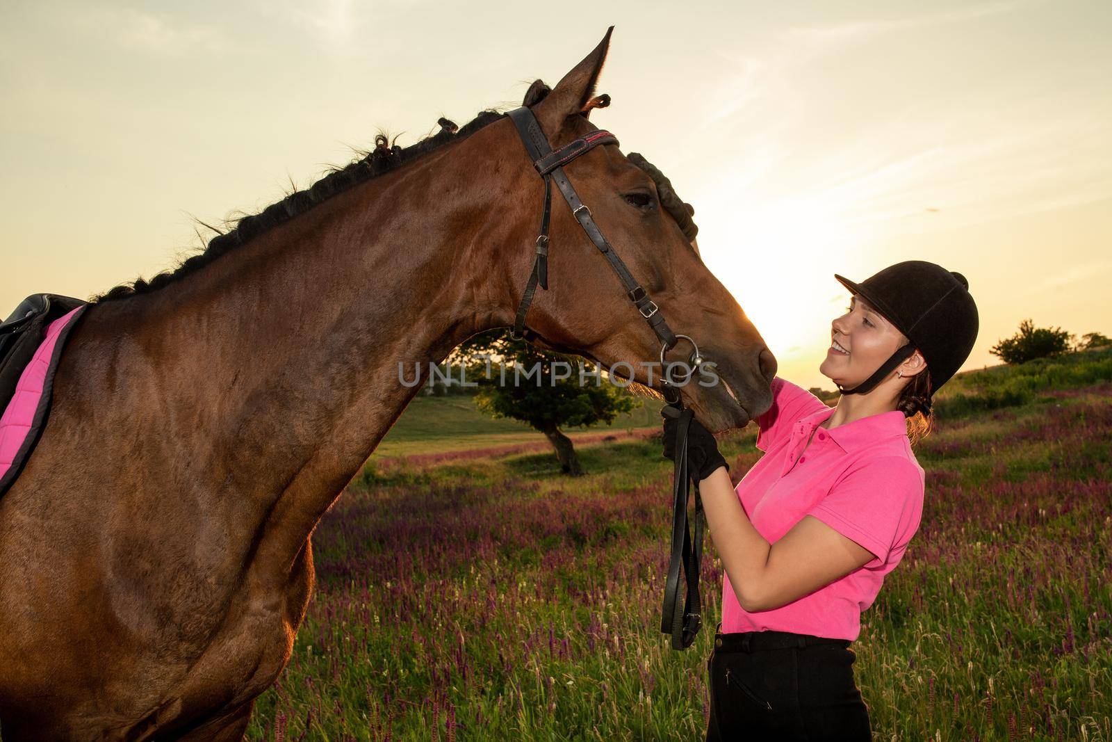Beautiful smiling girl jockey stand next to her brown horse wearing special uniform on a sky and green field background on a sunset. Equitation sport competition and activity.