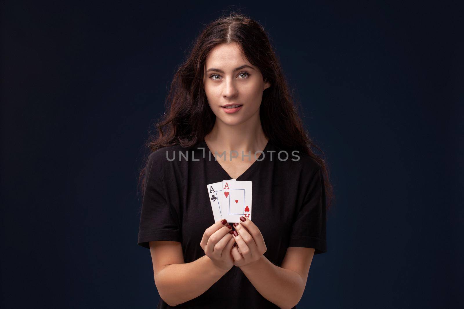 Curly hair brunette wearing a blackish t-shirt is posing with playing cards in her hands and looking at the camera. Poker concept on a black background. Casino.