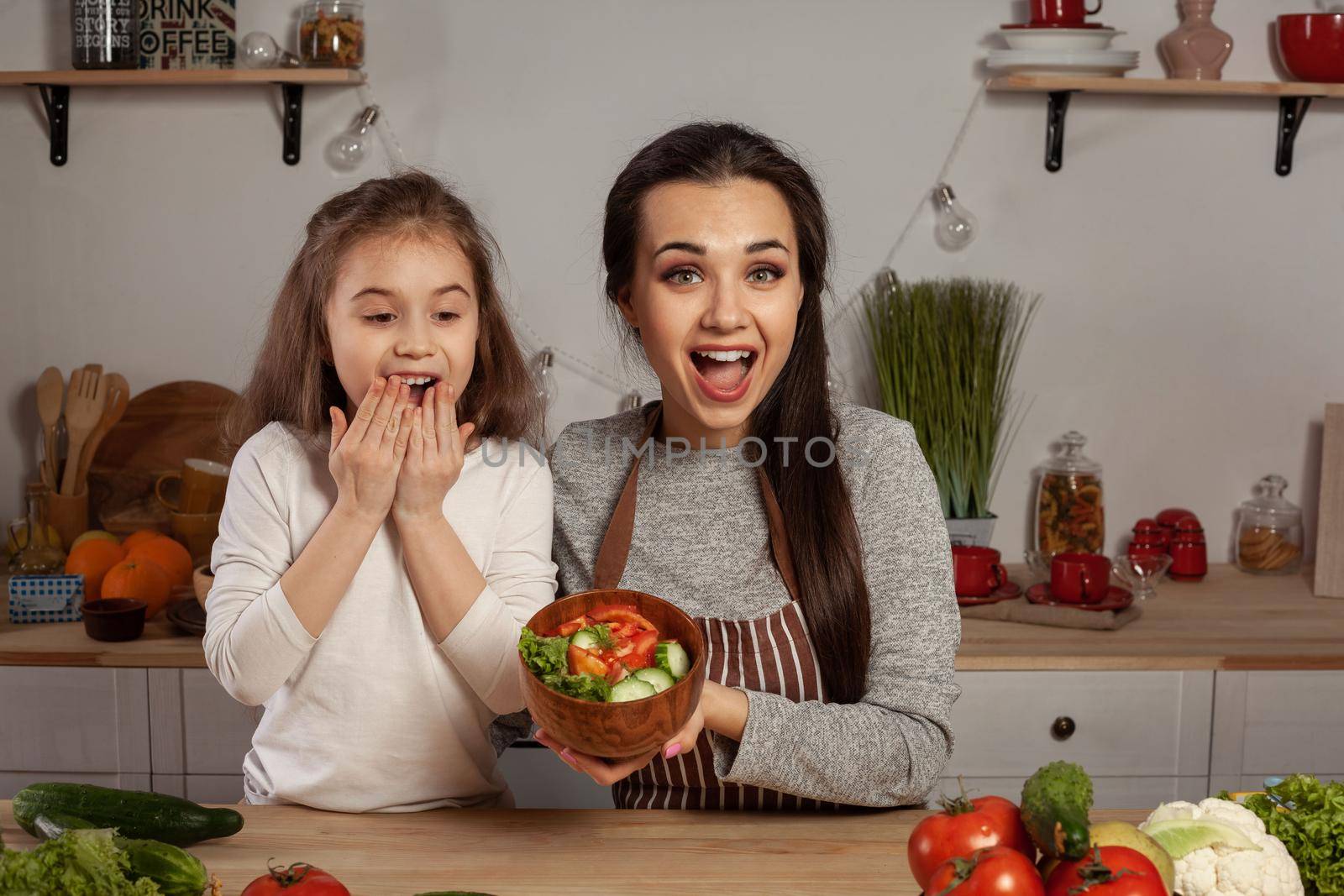 Happy loving family are cooking together. Smiling mommy and her little girl are making a vegetable salad and having fun at the kitchen, against a white wall with shelves and bulbs on it. Homemade food and little helper.