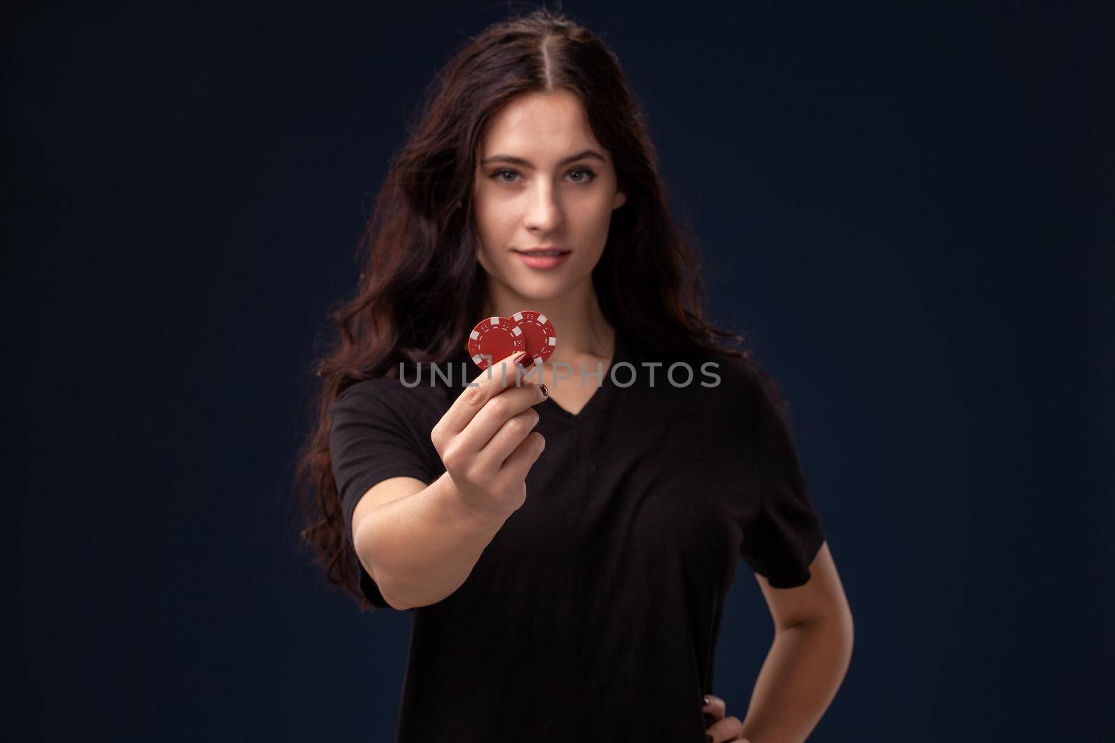 Beautiful woman wearing a blackish t-shirt is showing red gambling chips in her hands and looking at the camera. Poker concept on a black background. Casino.