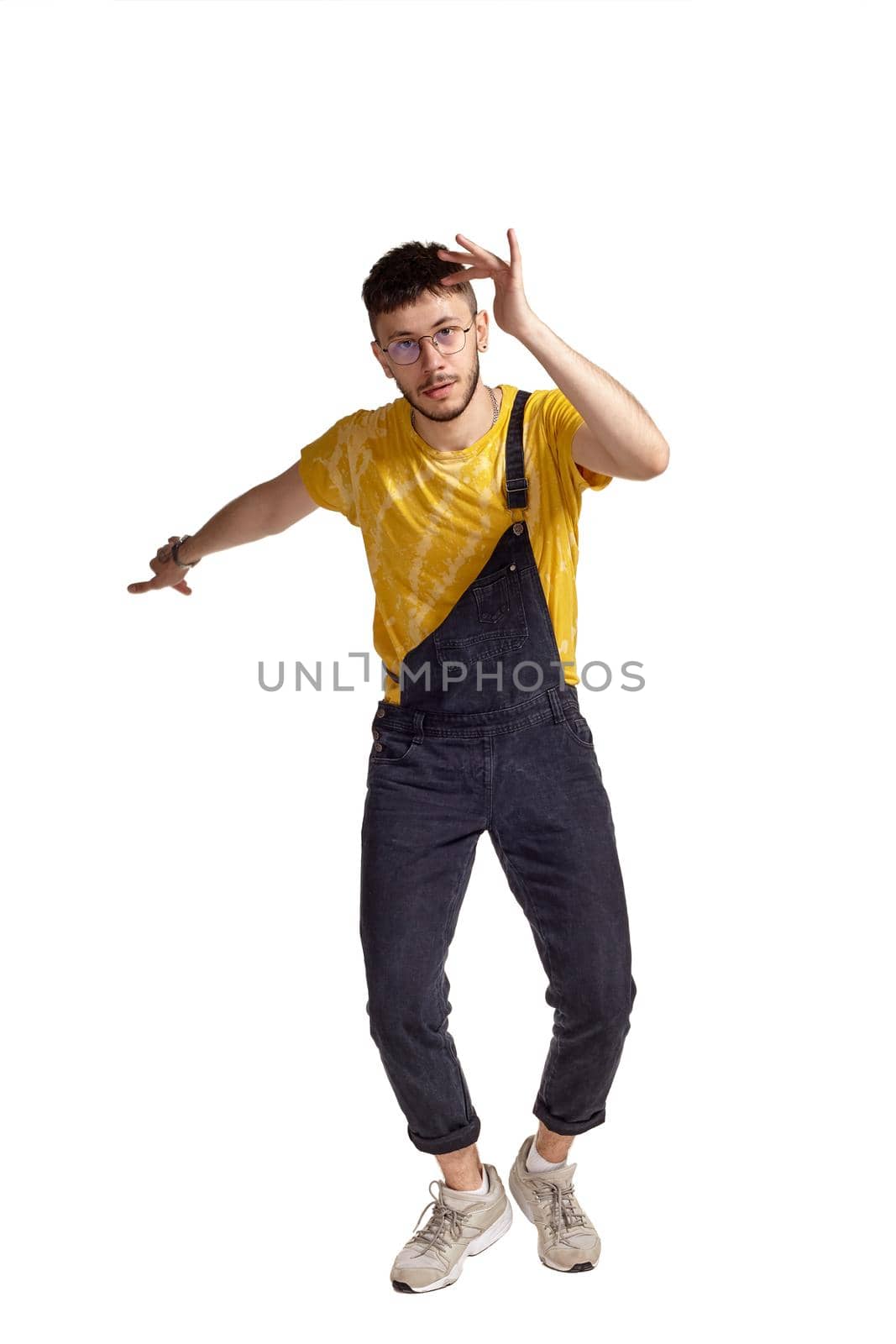 Full-length portrait of a fasionable guy in glasses, black jumpsuit, yellow t-shirt and gray sneakers fooling around in studio. Indoor photo of a man making dancing elements isolated on white background. Music and imagination concept.