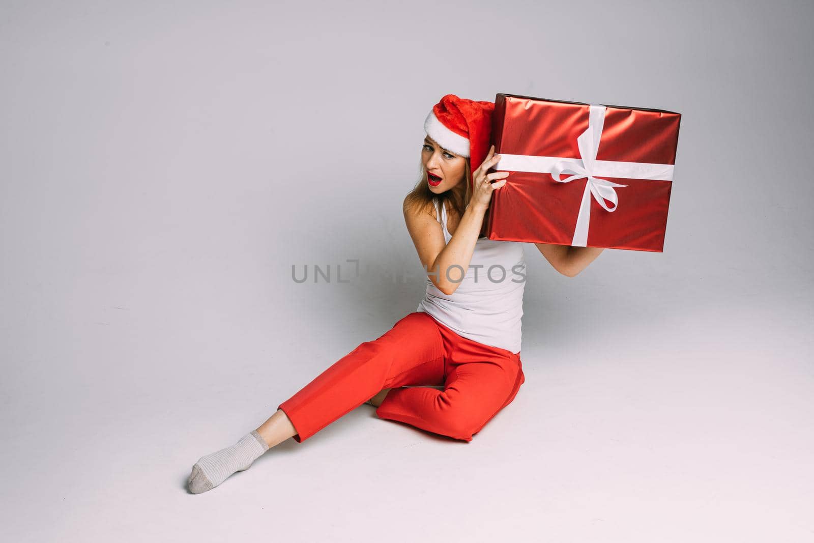 funny woman with christmas hat sits with a big red box and try to hear what is inside, picture isolated on white background by StudioLucky