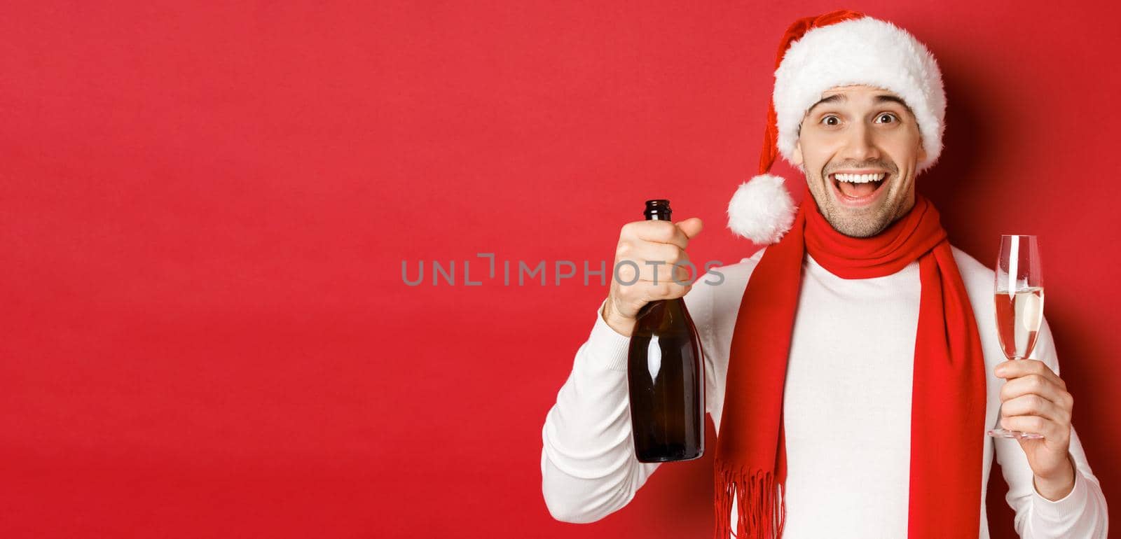 Concept of winter holidays, christmas and lifestyle. Close-up of cheerful handsome man, holding champagne bottle and glass, celebrating new year, standing over red background.