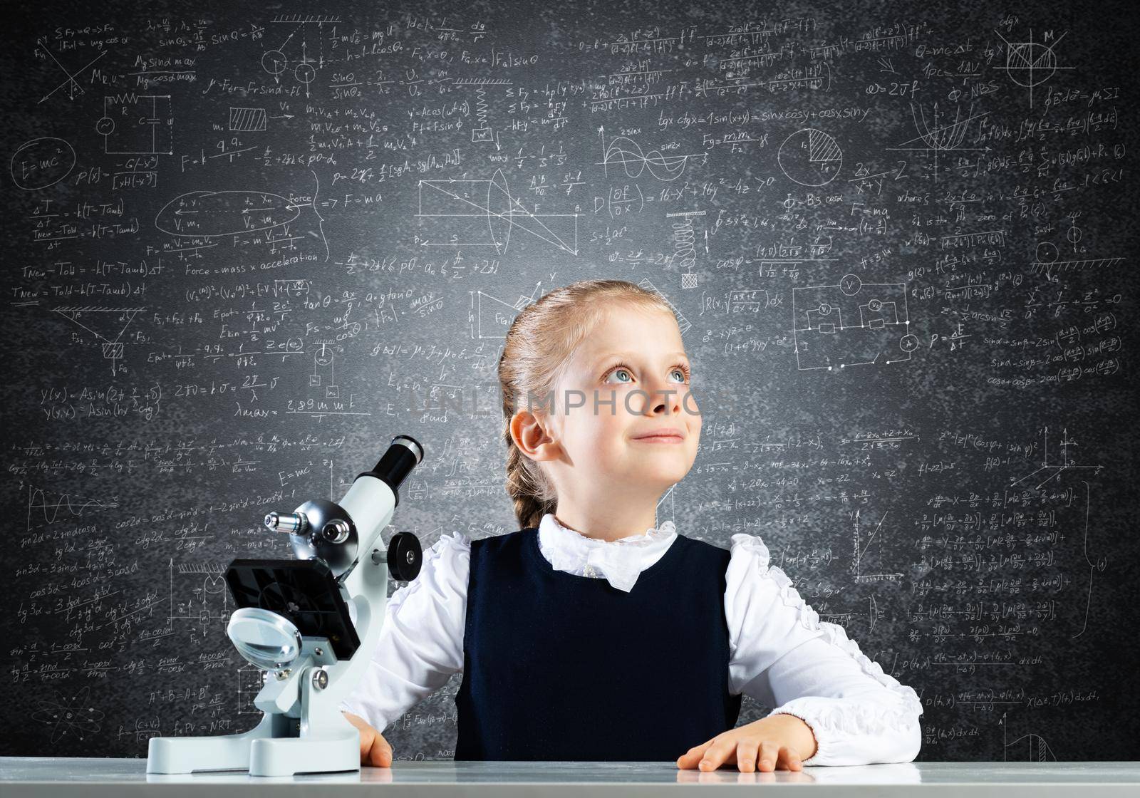 Little girl scientist with microscope on chalkboard background. Research and discovery concept with copy space. Elementary science class in modern school. Schoolgirl in schoolwear sitting at desk.
