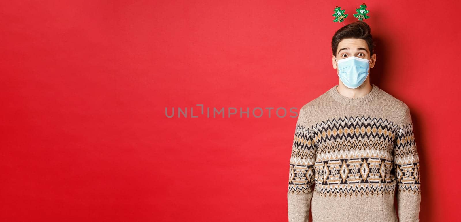 Concept of christmas, covid-19 and social distancing. Cheerful young man in medical mask and new year clothing, celebrating holidays during pandemic, standing over red background.