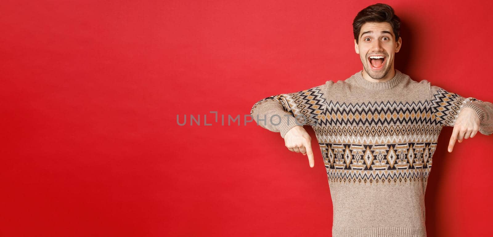 Concept of christmas celebration, winter holidays and lifestyle. Excited caucasian male model in xmas sweater, pointing fingers down and making announcement, showing advertisement, red background.