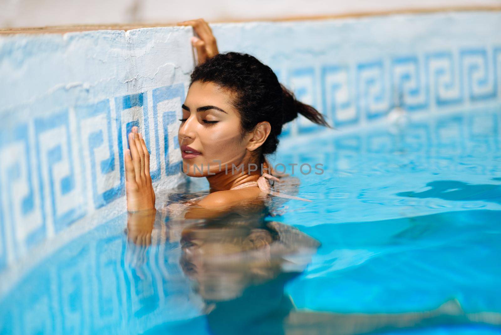 Beautiful Arab woman relaxing in swimming pool with eyes closed. Girl with healthy tanned skin and wet hair enjoying Summer Sun.