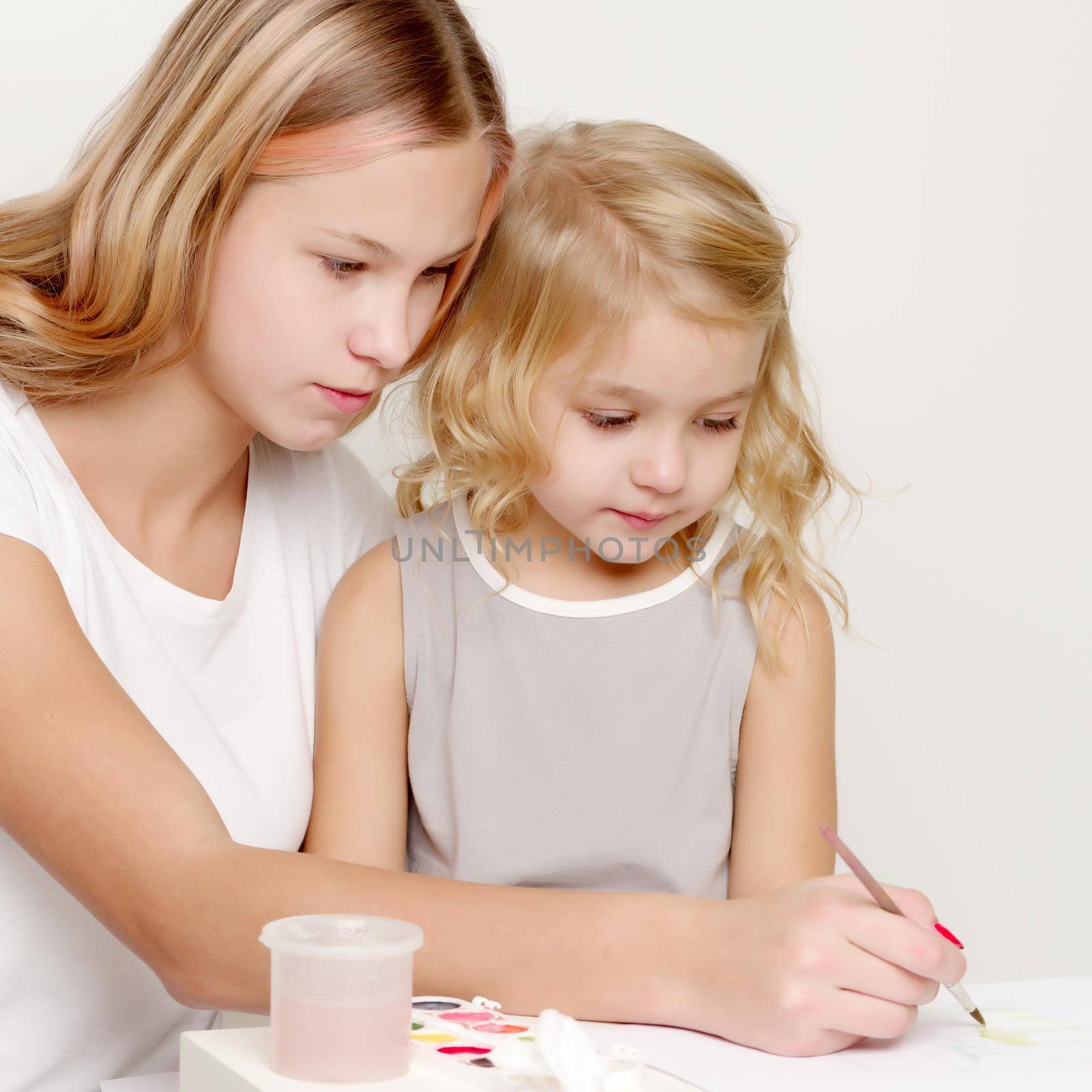 The older sister paints with the younger sister paints at the table on a piece of paper.