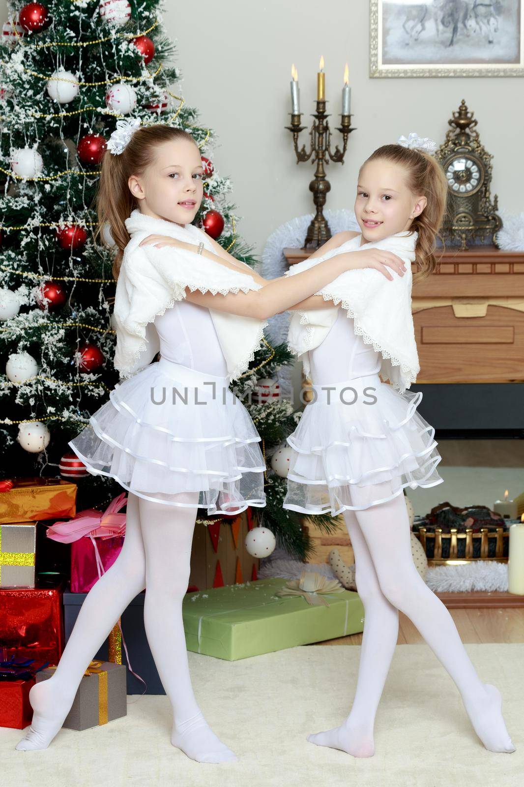 Little twin girls in beautiful white dresses hugging near the fireplace which burn Christmas candles.