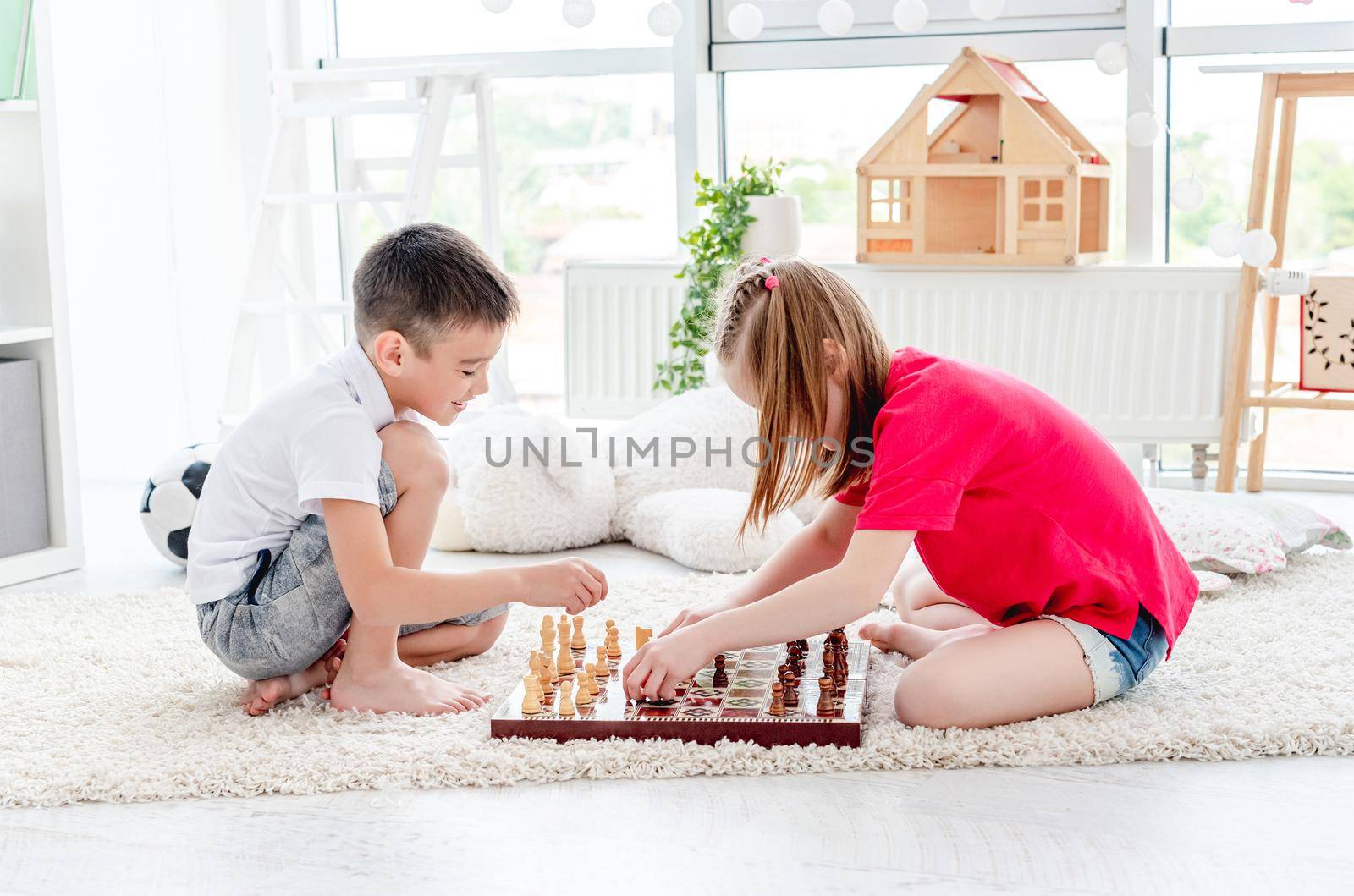 Cute boy and girl children playing chess sitting on floor in light room
