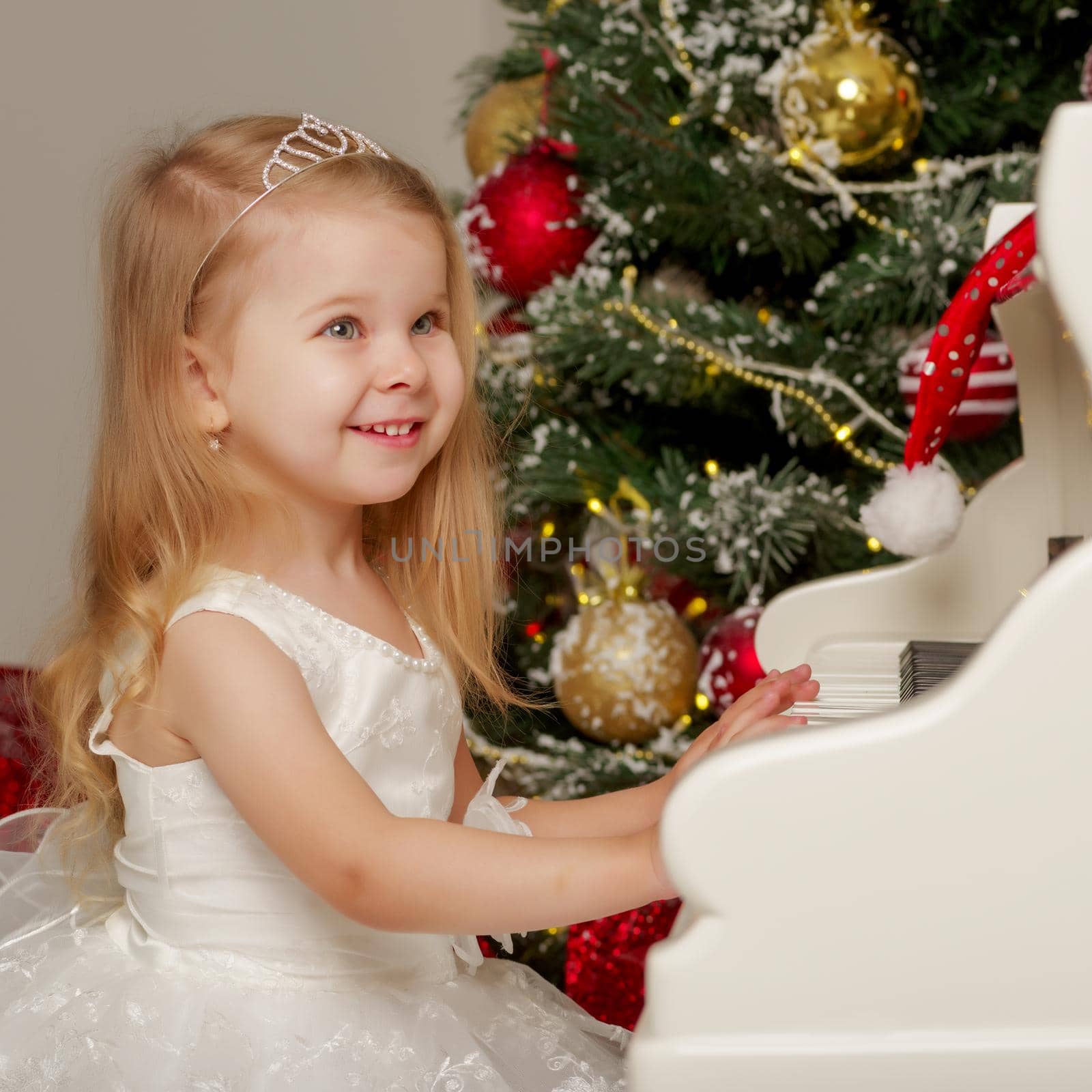 A cute little girl sitting near a white piano and a Christmas tree. The concept of the New Year, family holidays.