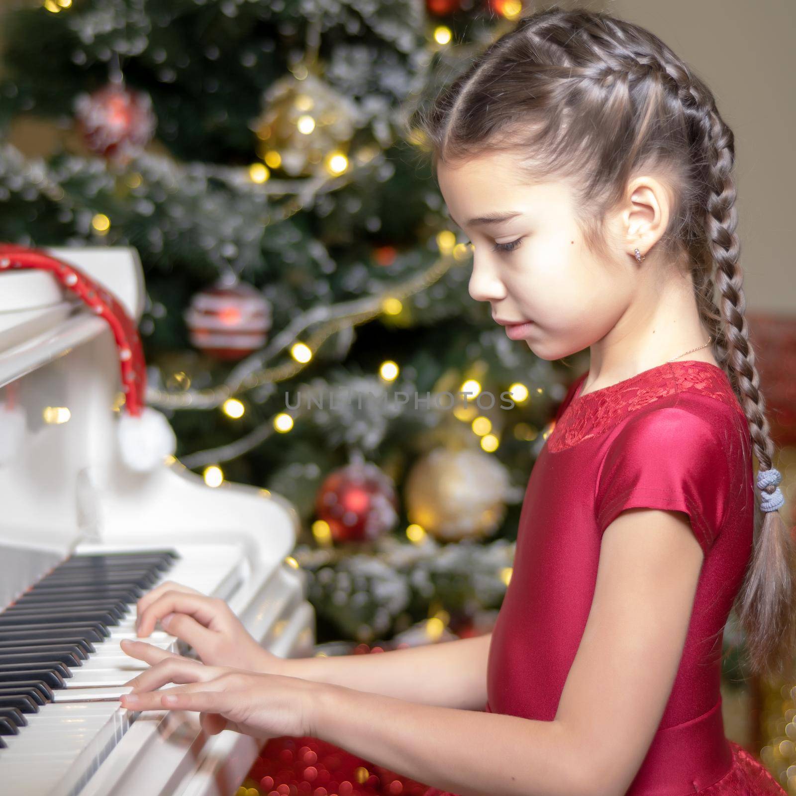 A cute little girl sitting near a white piano and a Christmas tree. The concept of the New Year, family holidays.