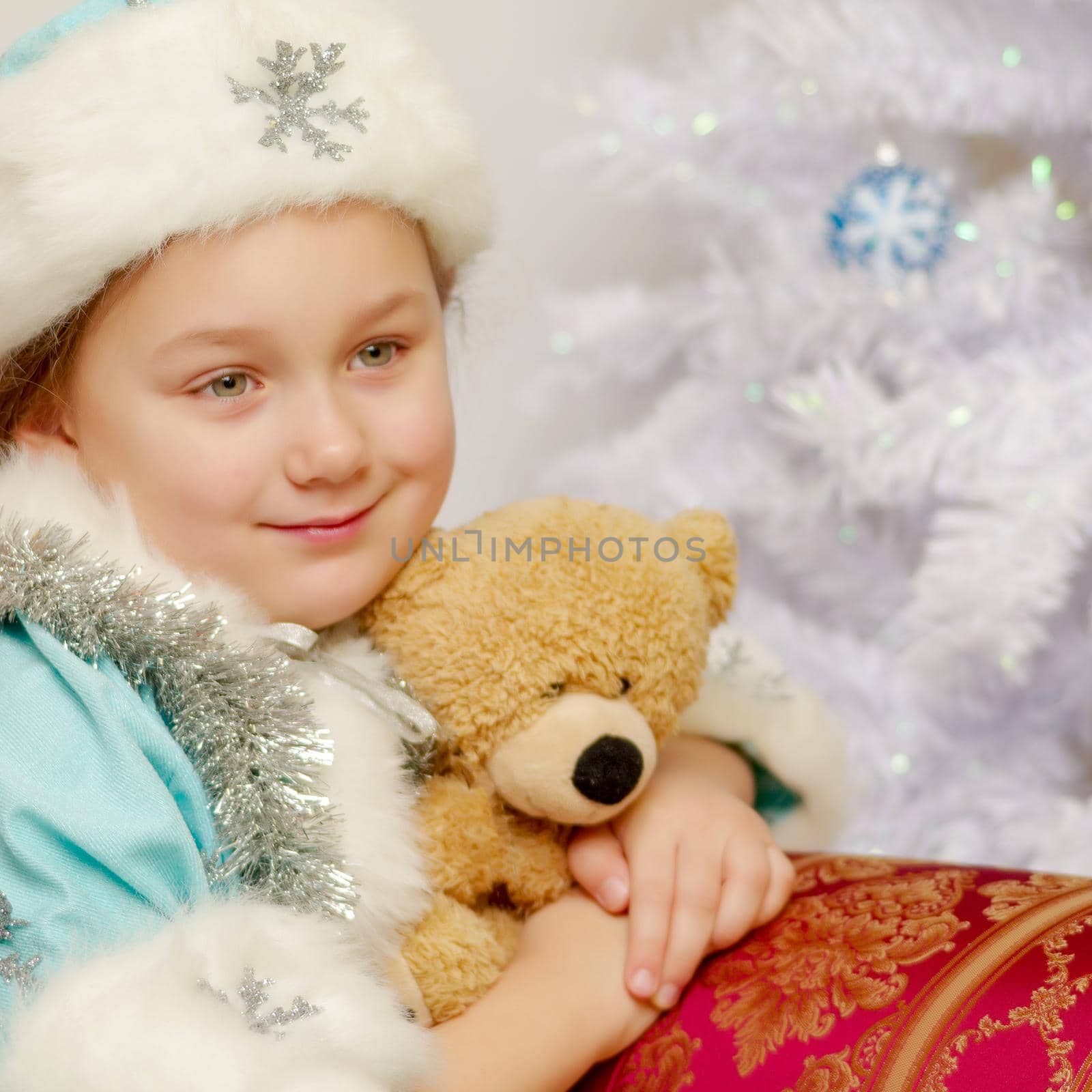 Cute little girl in the costume of the Snow Maiden near the Christmas tree, with a teddy bear. The concept of holidays.