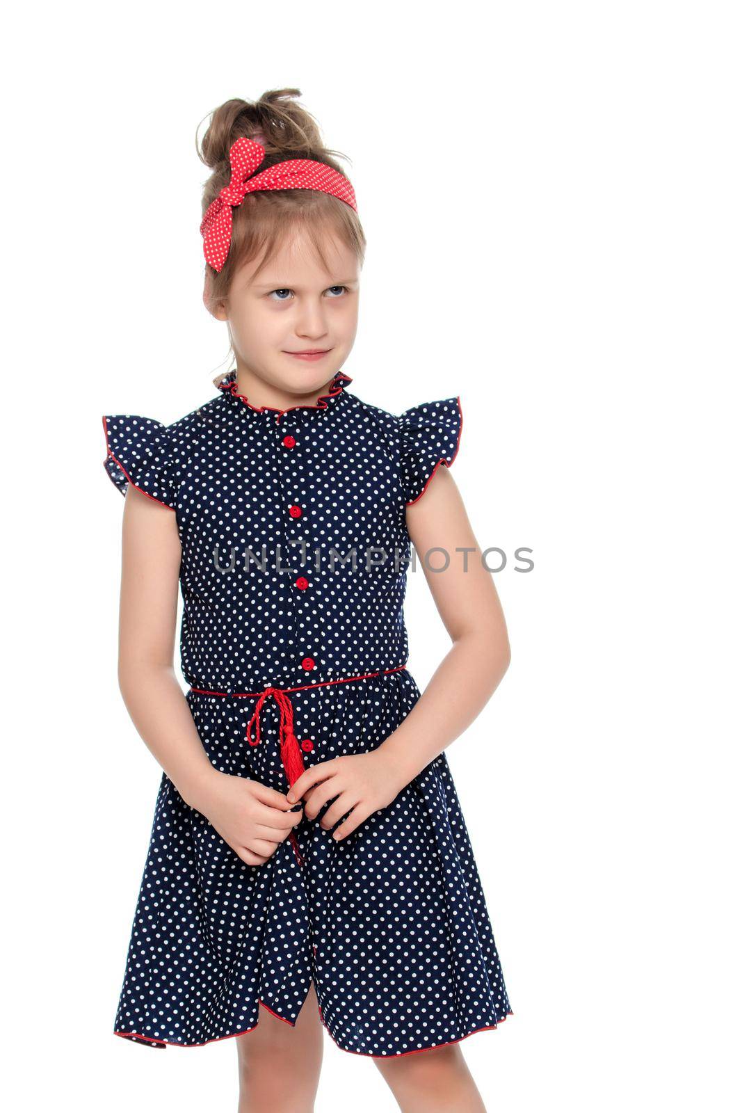 Little girl in blue polka dot dress with a red bow on his head.She shyly pulls hands to the edge of the dress.Isolated on white background.