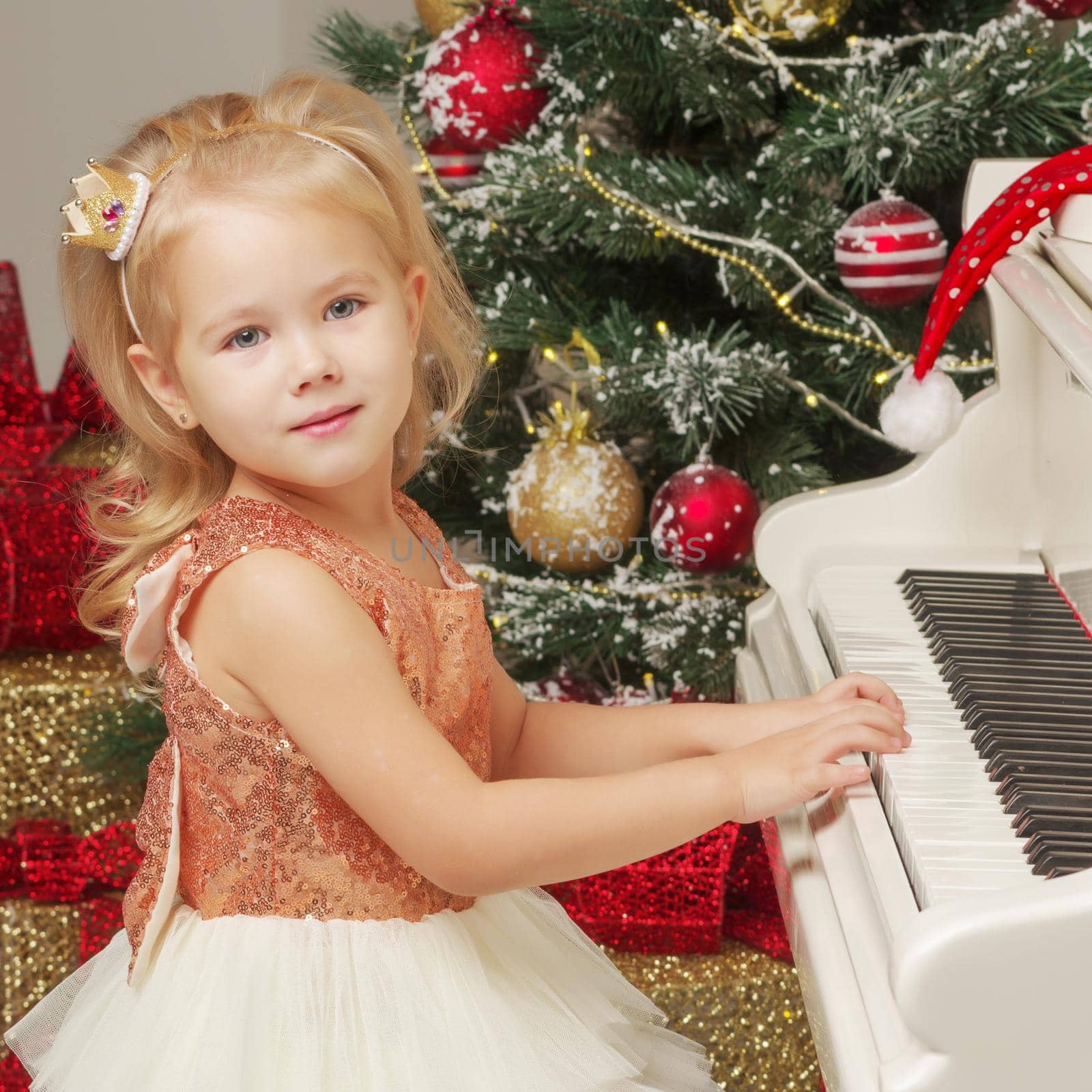 A cute little girl sitting near a white piano and a Christmas tree. The concept of the New Year, family holidays.