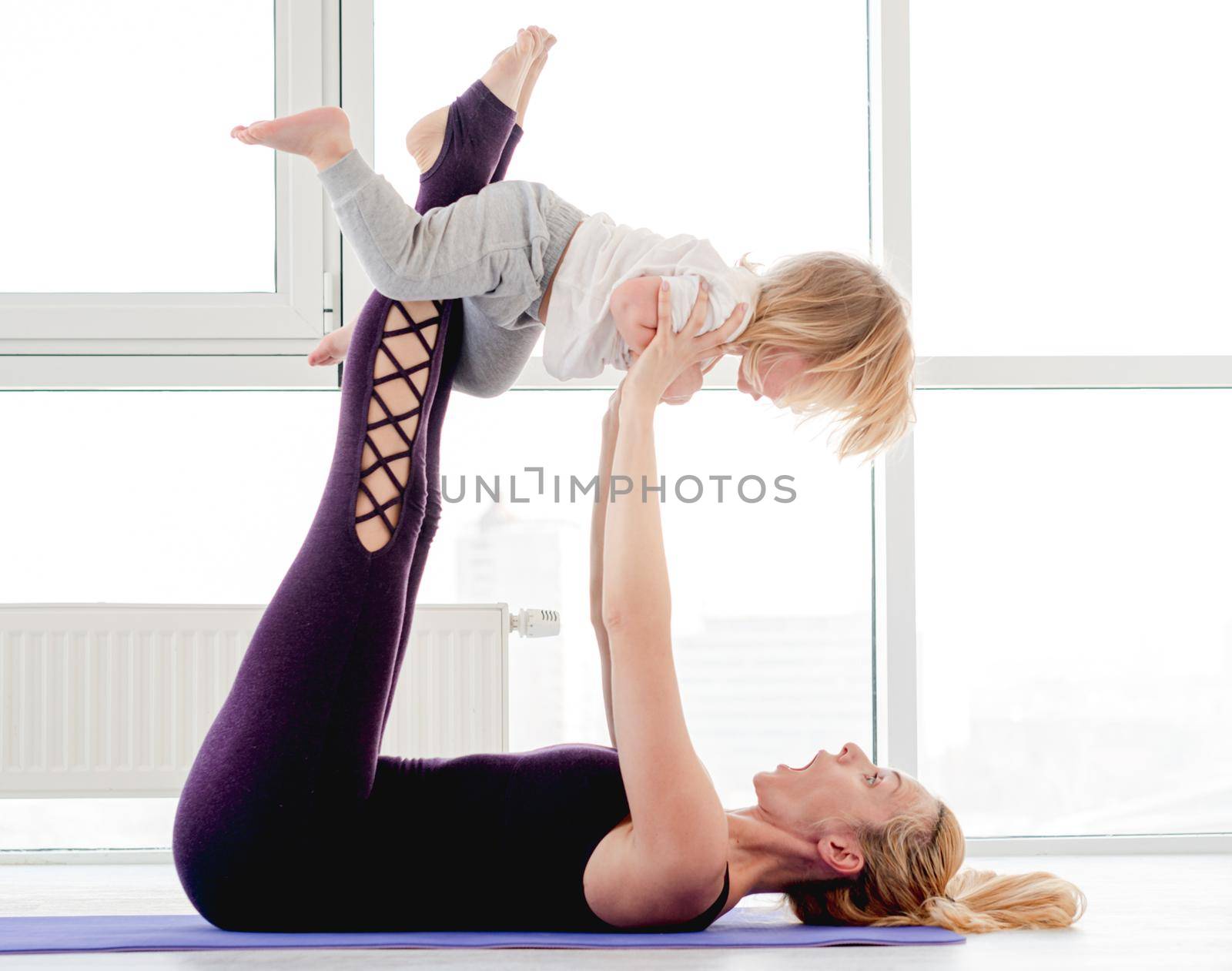 Young blond girl mother practicing yoga with child and holds her son in outstretched arms. Family partner stretching workout