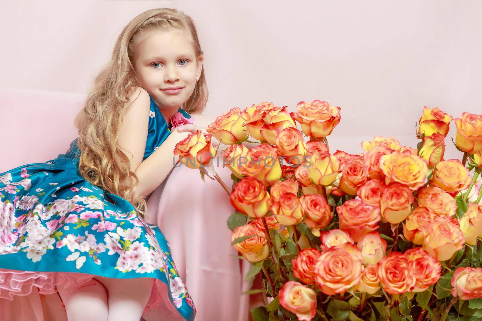 A beautiful little girl with long, light, curly hair, in a blue long skirt. She sits on a pink couch beside a large bouquet of tea roses.