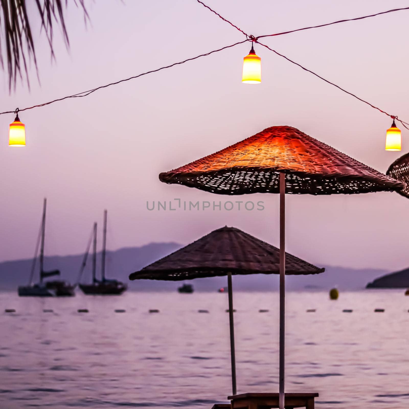 Beach umbrellas and lanterns against the backdrop of the sea, islands, mountains and yachts at sunset. Summer vacation concept
