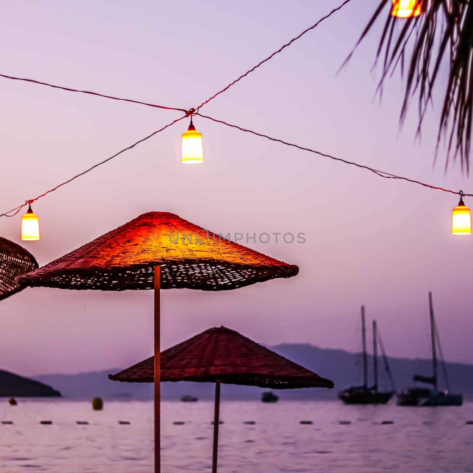Beach umbrellas and lanterns against the backdrop of the sea, islands, mountains and yachts at sunset. Summer vacation concept