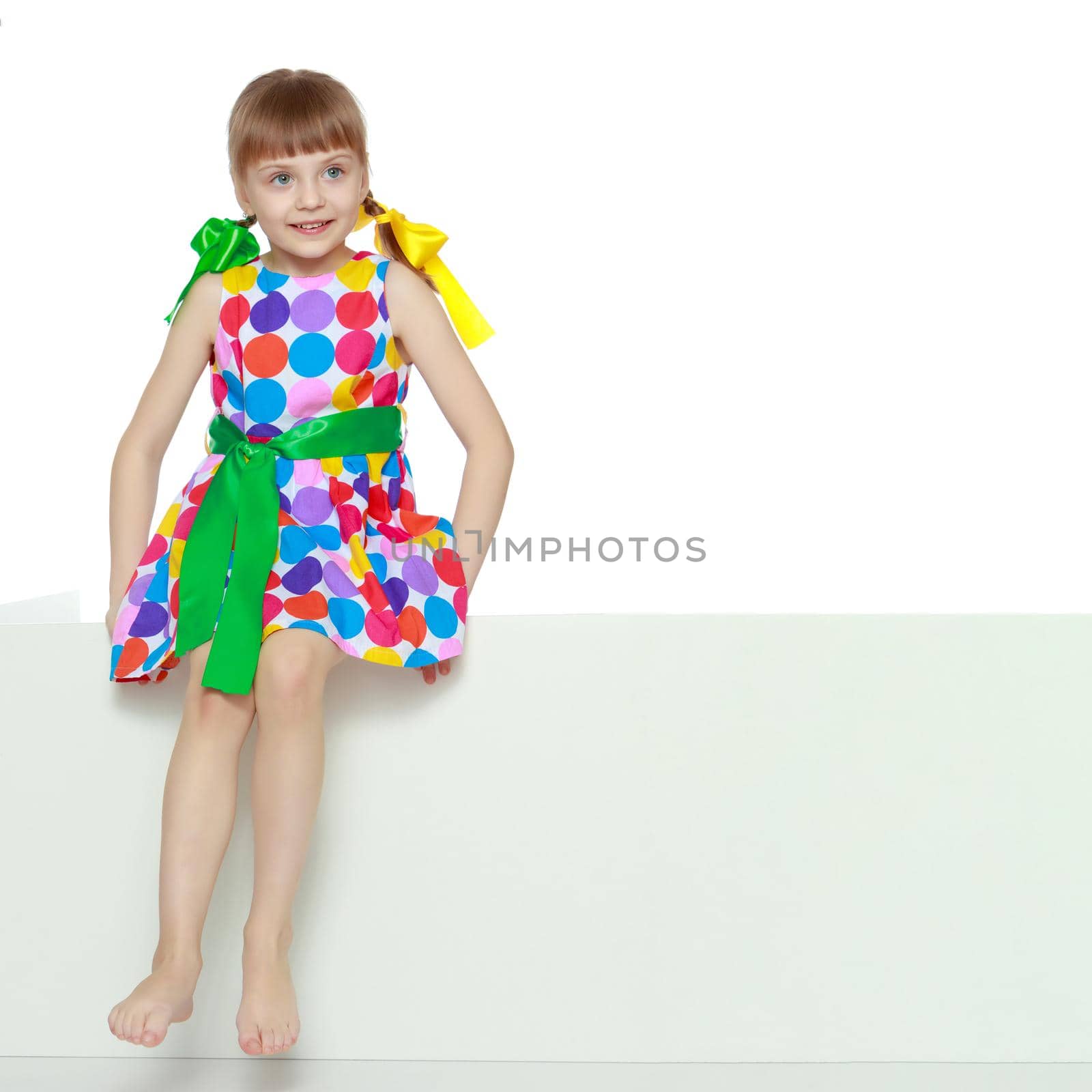 A beautiful Caucasian little blonde girl with long pigtails, in which large colored bows are braided, and a short bangs on her head. In a short summer dress.Looks because of the white obstacle.