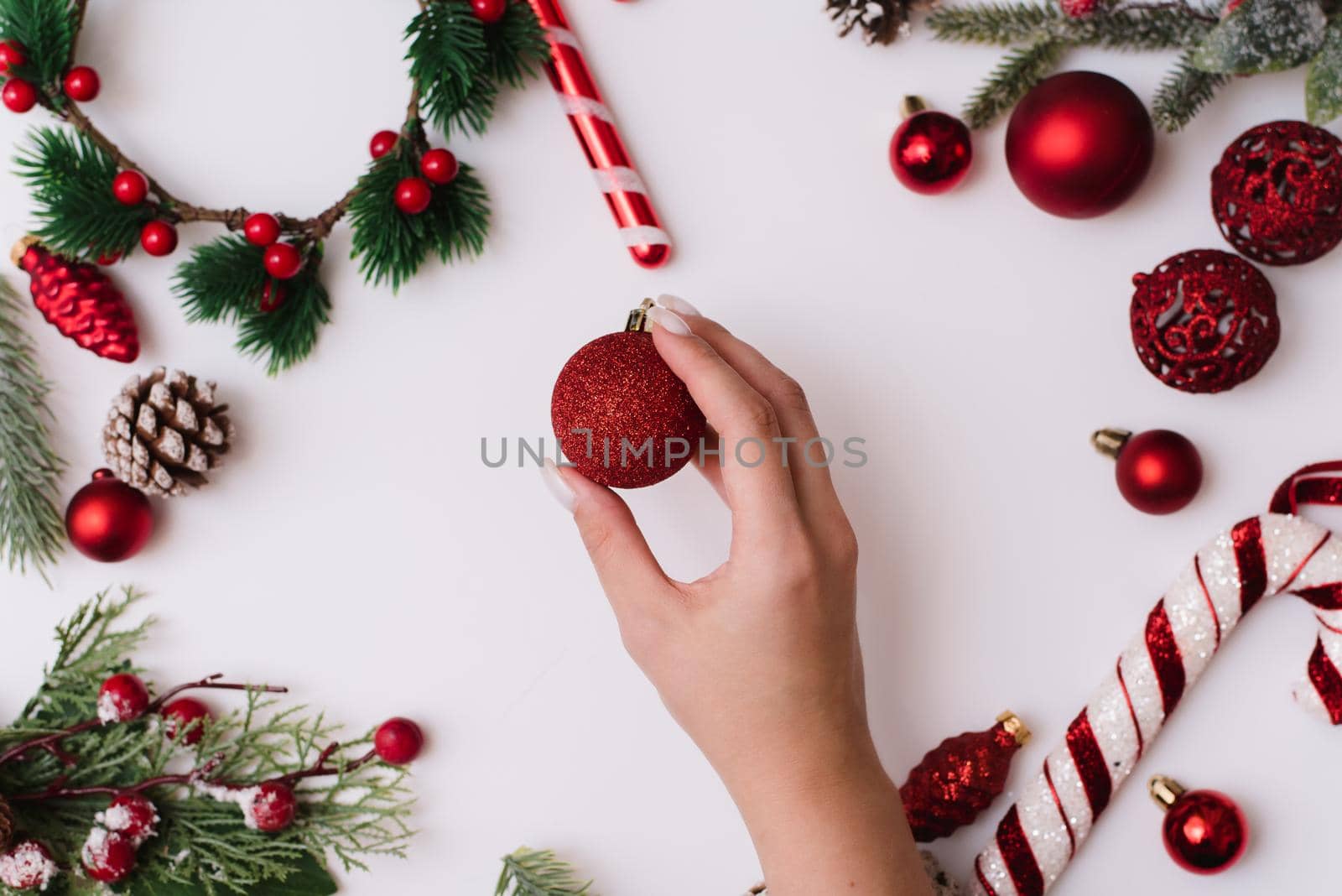 A female hand with a white manicure holds a red ball on a white background with Christmas branches and red decorations.