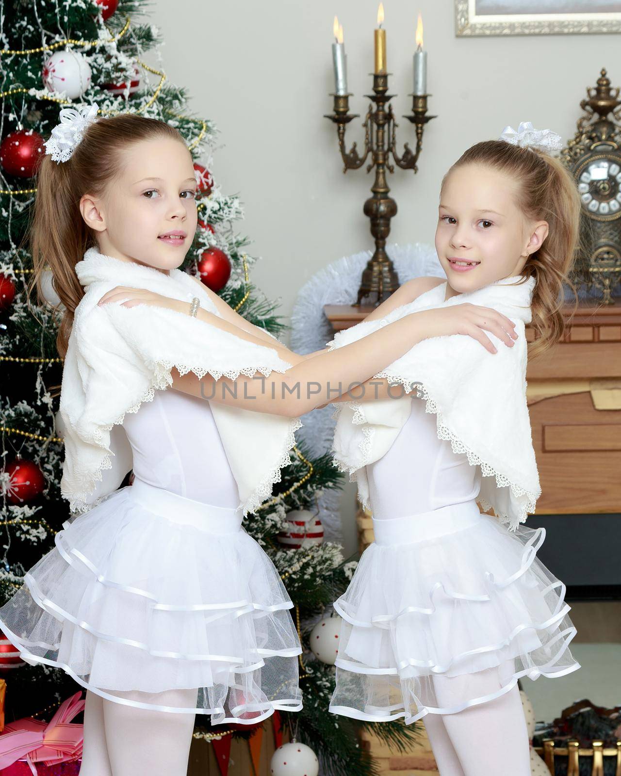 Little twin girls in beautiful white dresses hugging near the fireplace which burn Christmas candles.