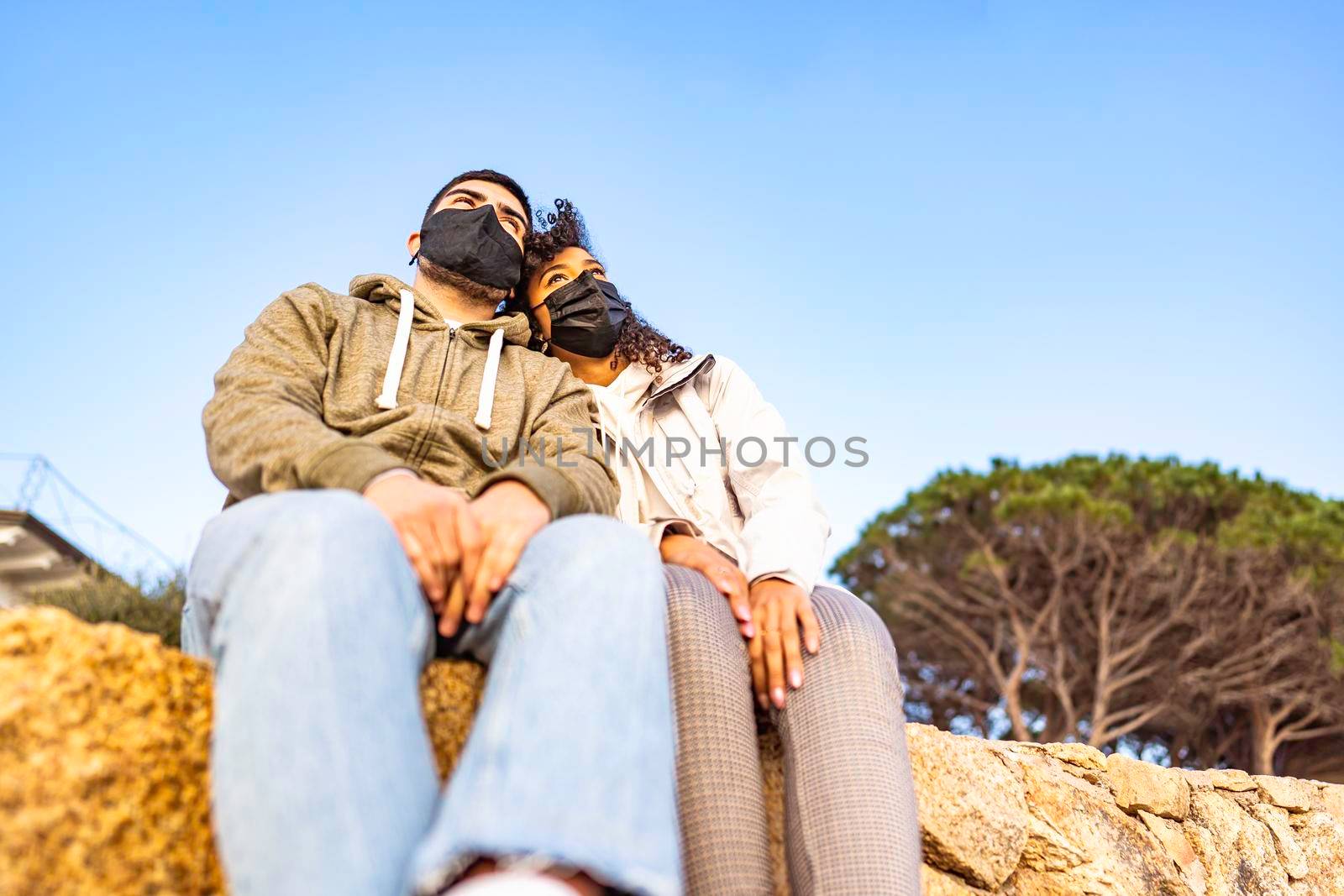 Young multiracial couple in love sitting on a wall wearing black safe protective face mask head to head looking at horizon at sunset. Pandemic global issue disturbing normal life and people future by robbyfontanesi