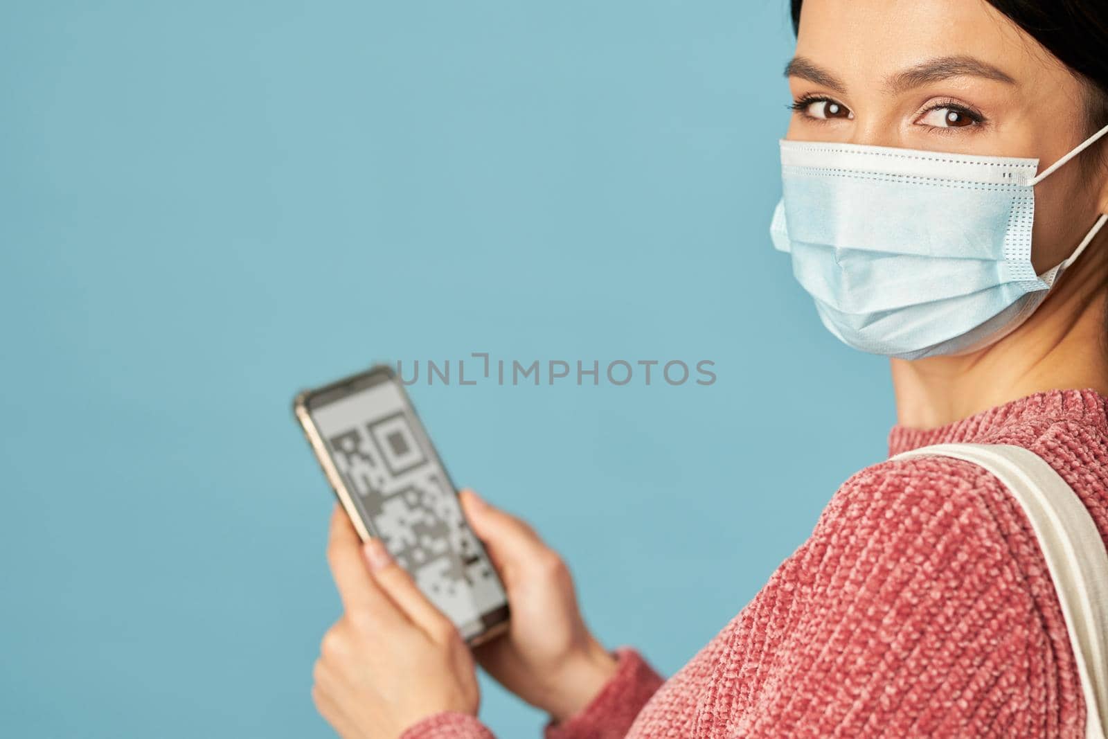 Cropped photo of happy woman in mask holding mobile phone while posing in studio, isolated on blue background. Copy space. Quarantine, coronavirus concept