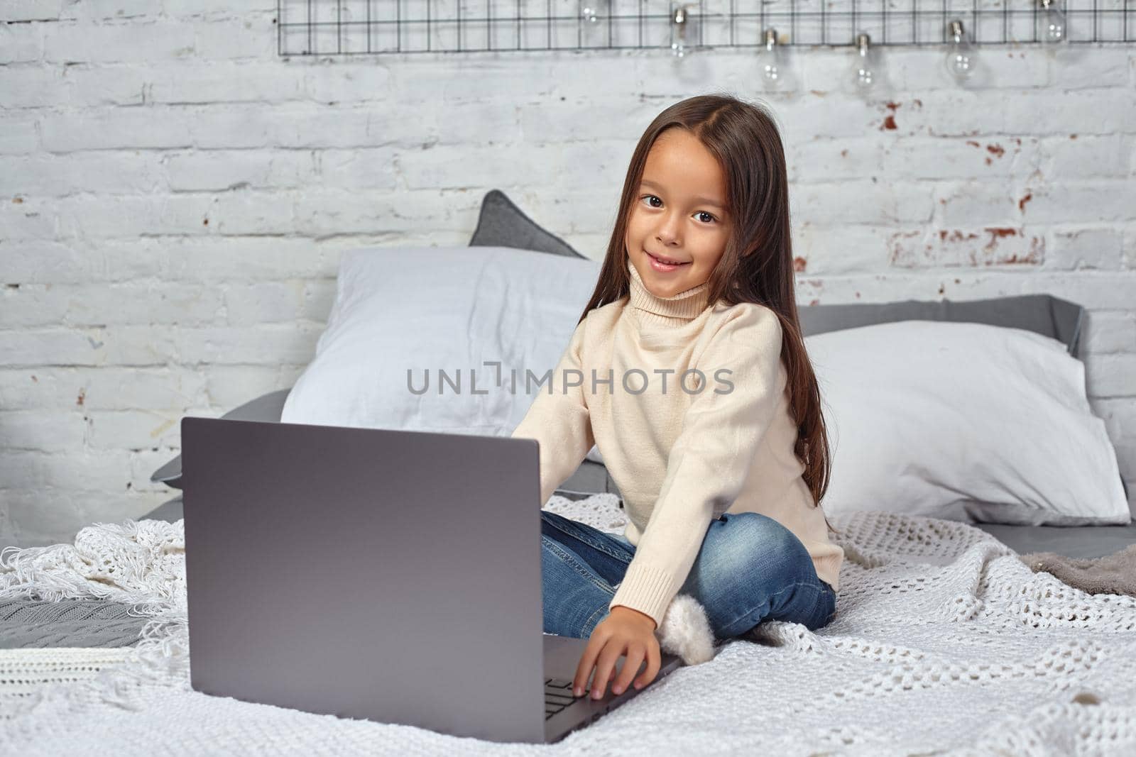 Cute little girl girl feeling amusing while watching cartoons on a laptop sitting on bed. White sweater and jeans.