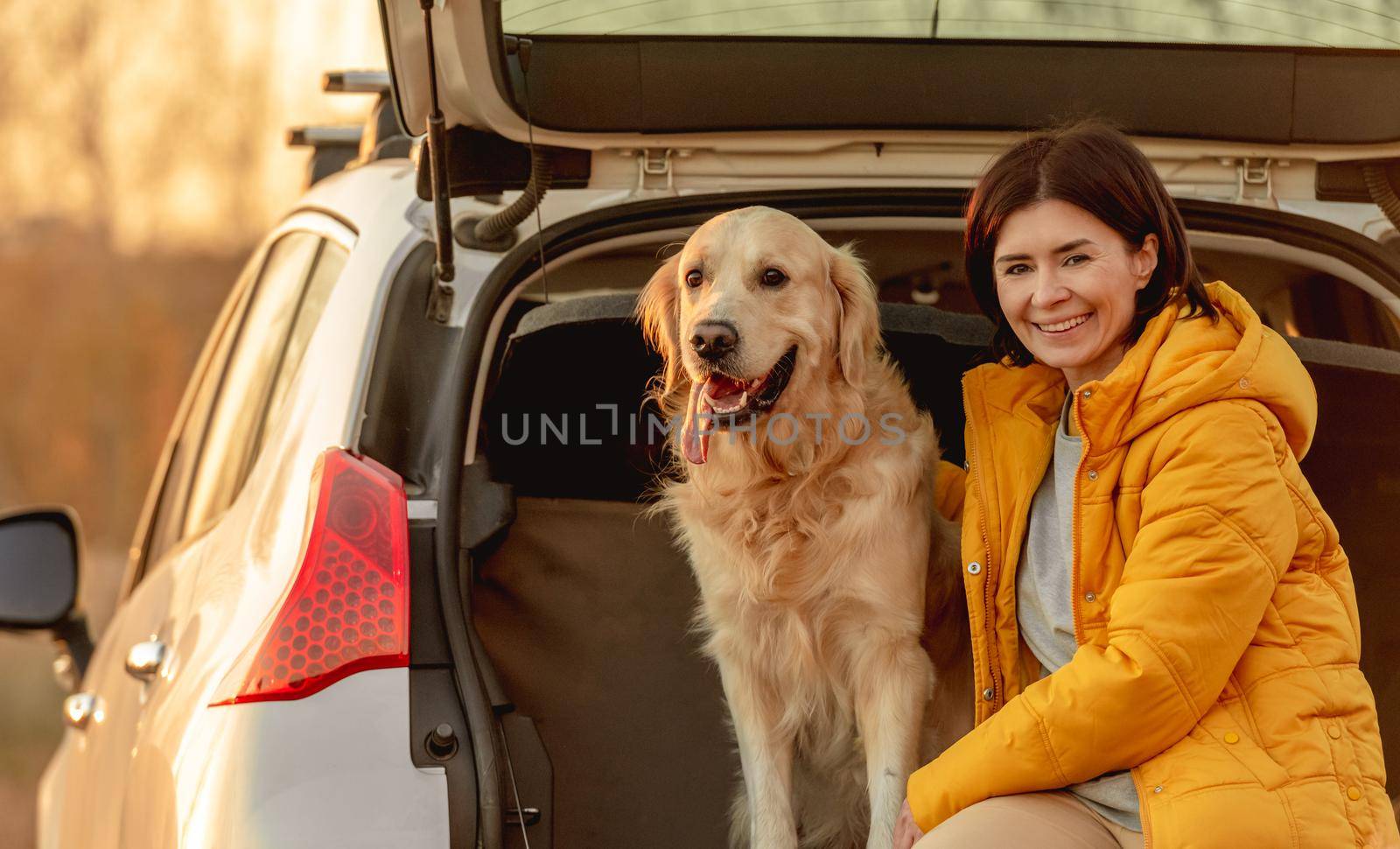 Girl with golden retriever dog sitting in car trunk outdoors. Young woman with doggy pet labrador in vehicle at nature at autumn