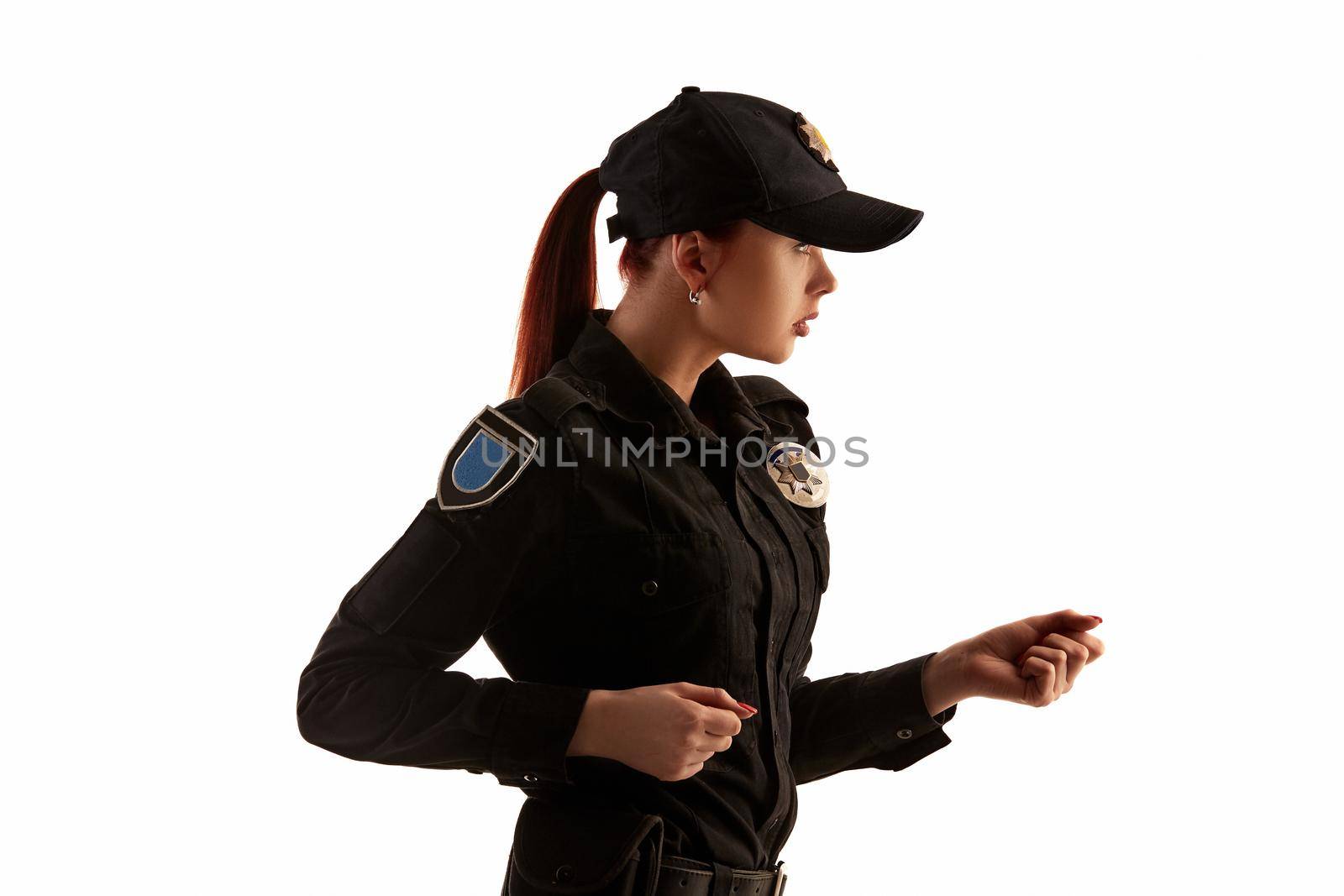Close-up shot of a nice redheaded policewoman in a black uniform and a cap preparing to fight. She is standing sideways isolated on white background. Defender of citizens is ready to enforce a law and stop a crime.