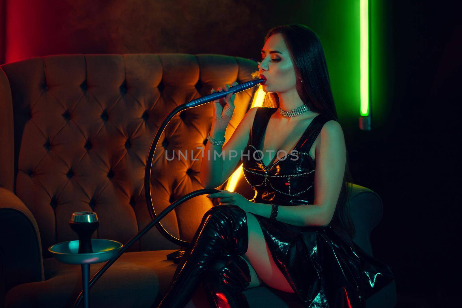Charming brunette woman with a perfect make-up, in a sparkling necklace, leather dress and patent boots is smoking a hookah sitting on a sofa at a luxury night club. Concept of pleasant spending time. Dark background with colorful neon backlights.
