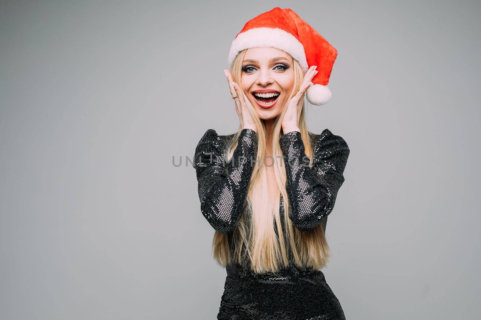 Stock photo of attractive jolly girl with long blonde hair in sparkling dark dress wearing red Santa hat holding hands at face in happiness.