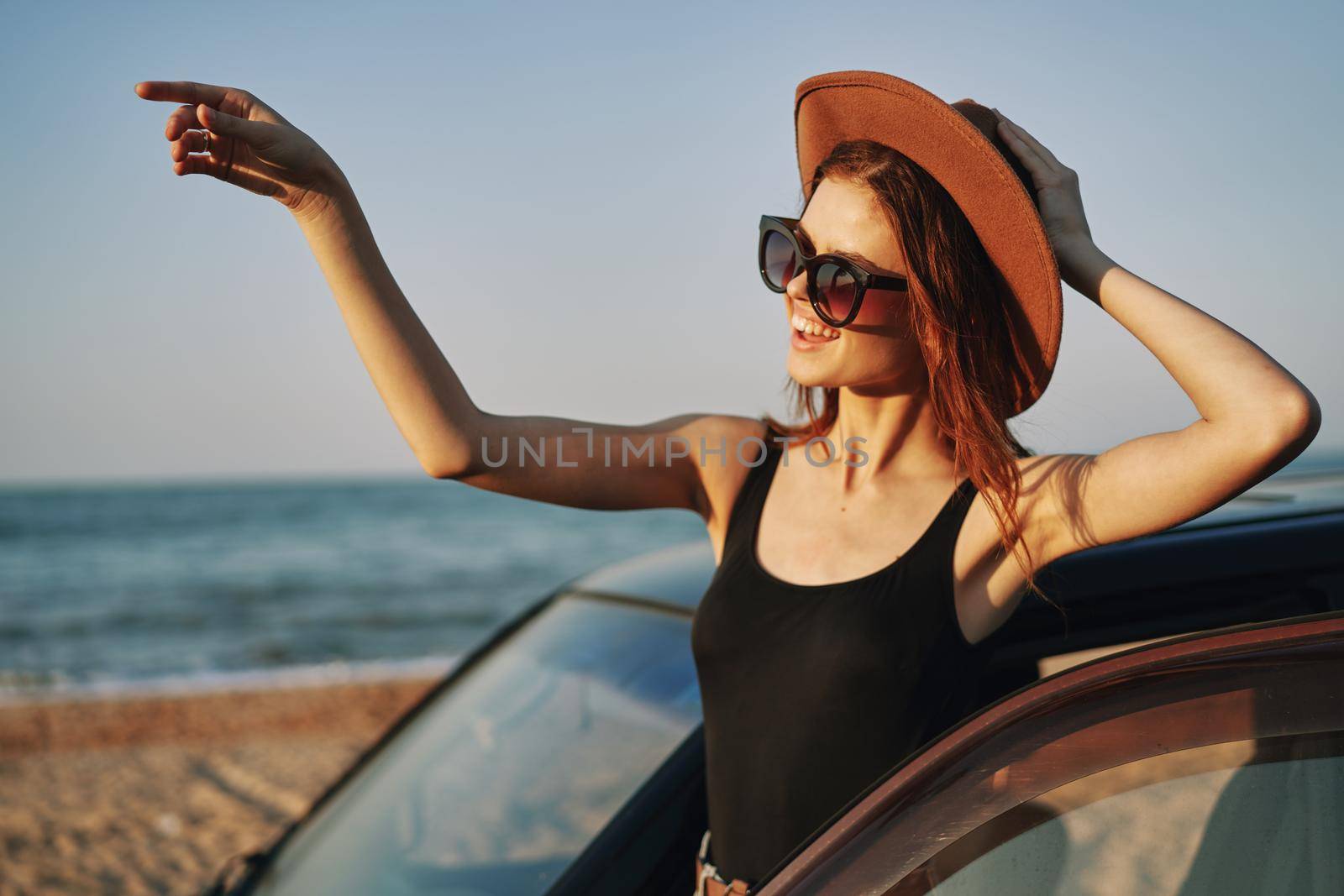 cheerful woman in sunglasses and a hat on the ocean near the car by Vichizh