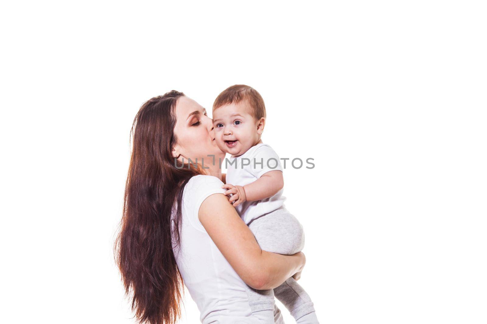 Beautiful young mother holds her child in her arms and kisses her baby, wearing white cloths, on a white background