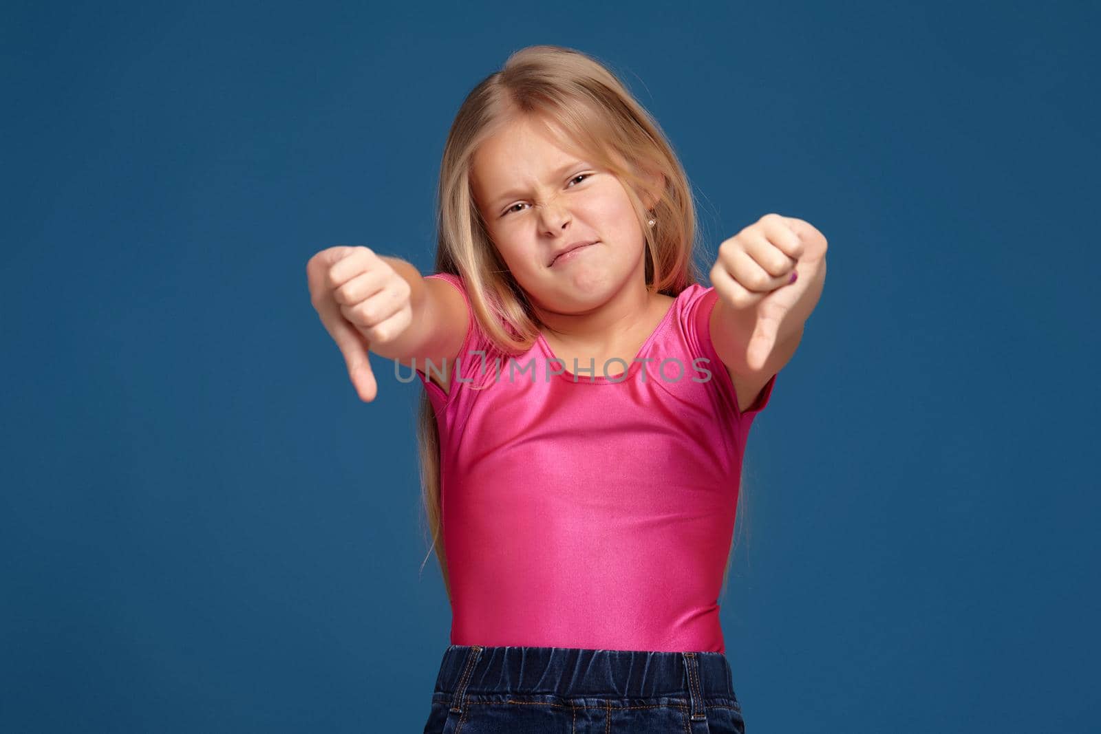 Close-up emotional portrait of caucasian little girl in pink shirt on blue background, making dislike, thumbs down gesture, looks unhappy