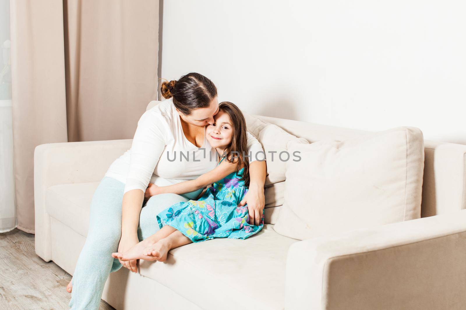 Mother and daughter at home. Mom sitting on the sofa gently hugs and kisses her smiling daughter.