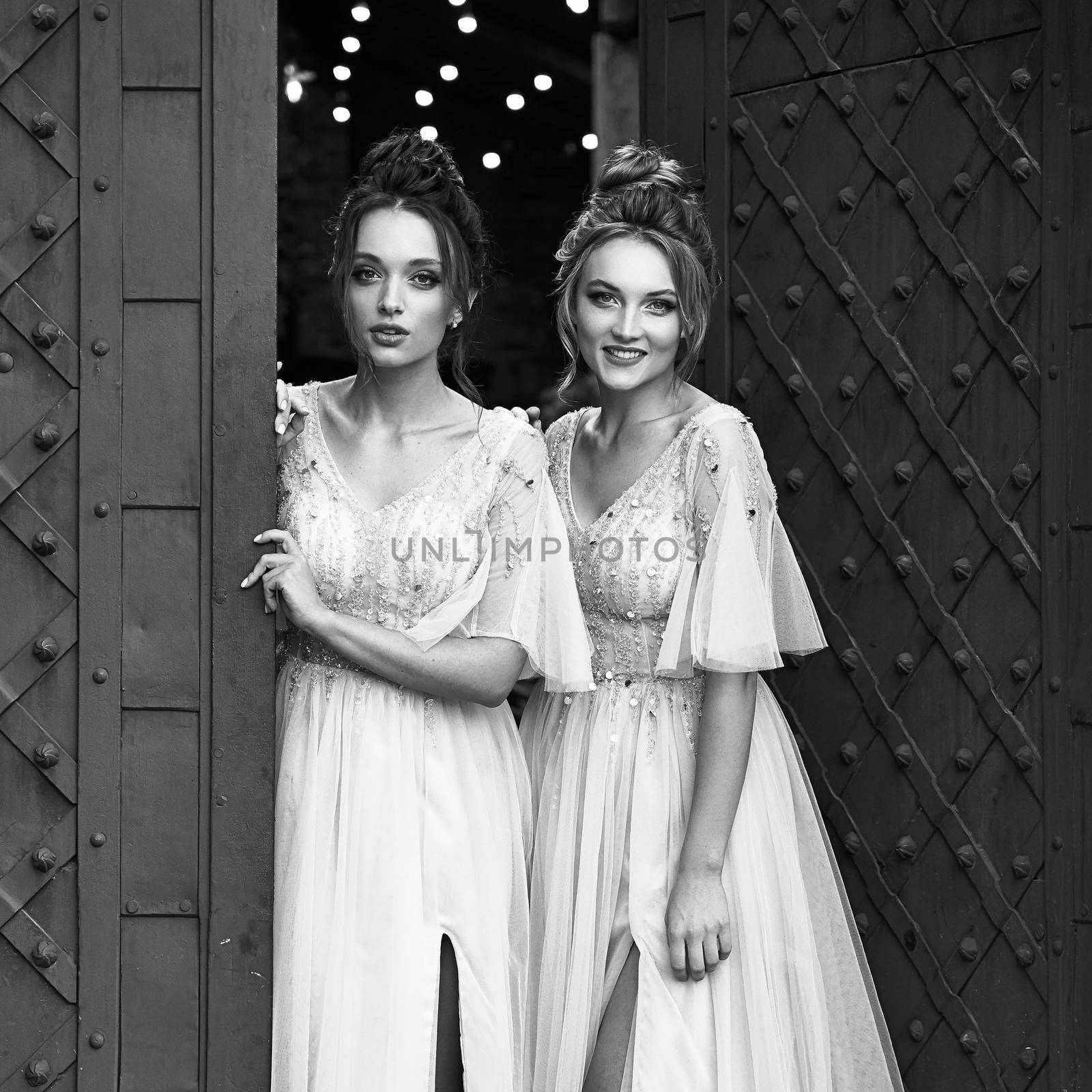 Beautiful retro styled bridesmaids ladies in gorgeous elegant stylish light grey silver floor length dresses in old beautiful European city welcome greeting guests at the gate. Black and white. by berezko