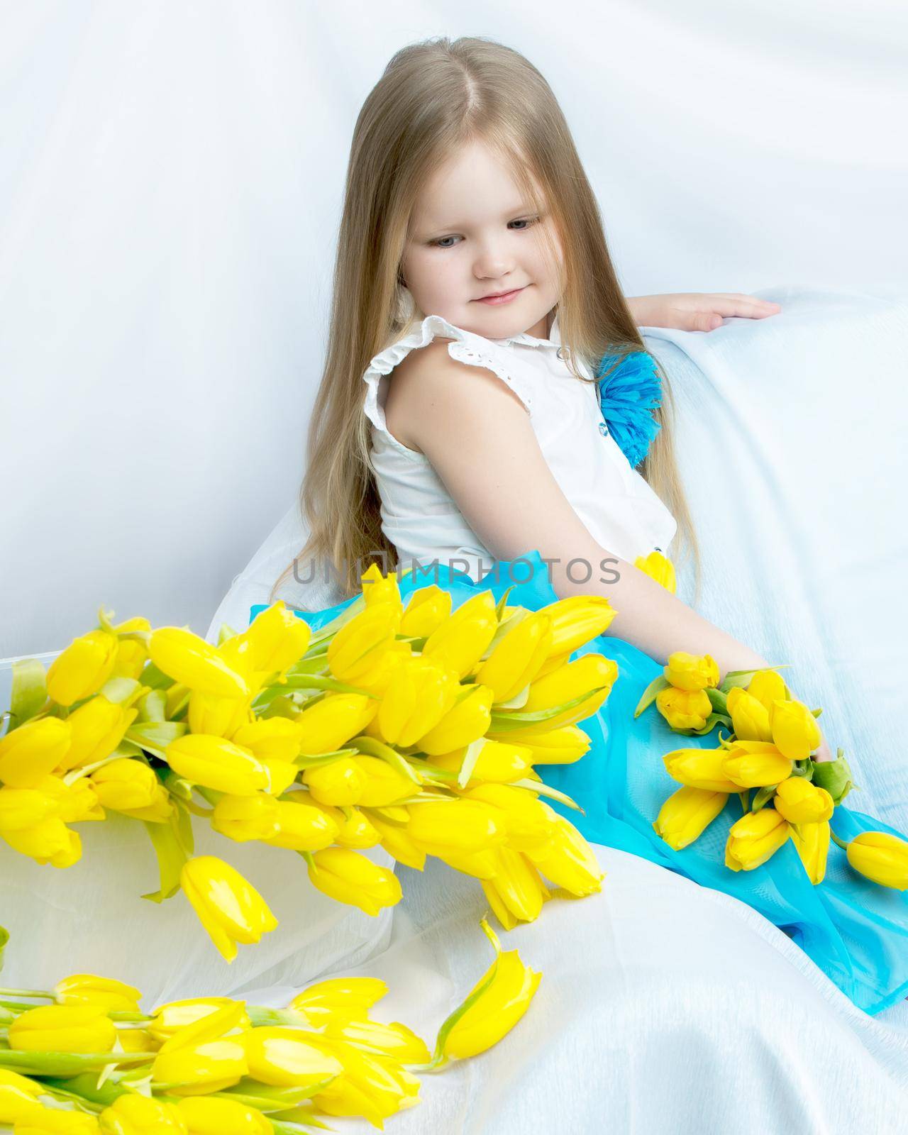 Cute little girl with long blond hair in a long blue skirt sitting on the couch with a large bouquet of bright yellow tulips.