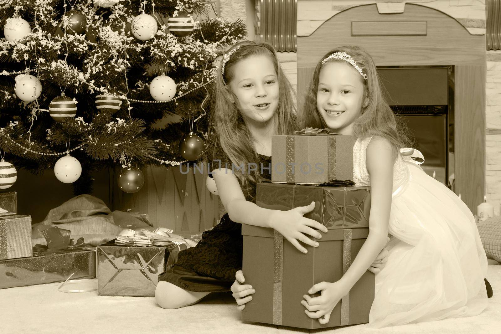 Cute little twin girls, sitting on the floor near the Christmas tree and electric fireplace on which candles are burning. In the hands of the girls boxes with gifts.Black-and-white photo. Retro style.