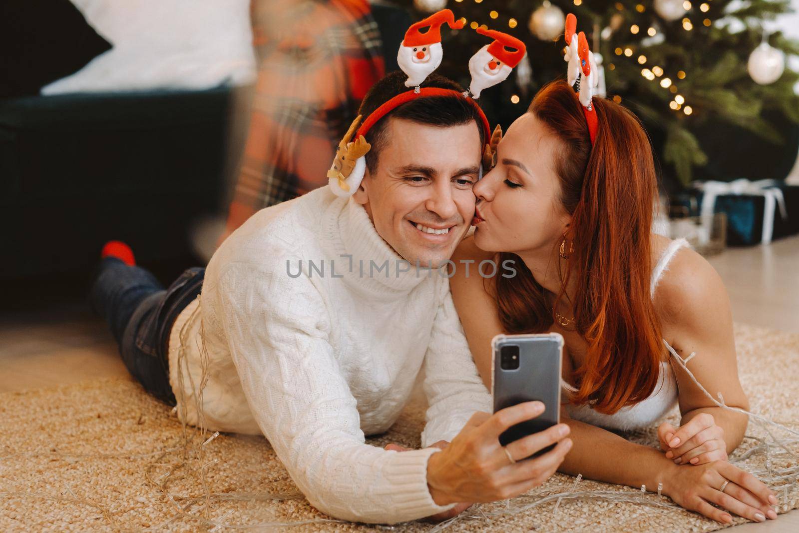 a happy married couple is lying on the floor at home near the Christmas tree and taking pictures of themselves.