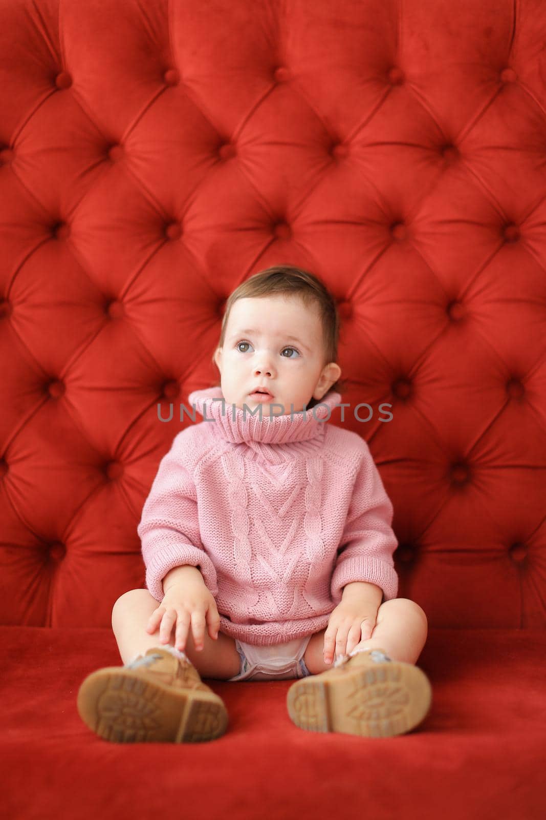 Little beautiful girl wearing pink sweater sitting on sofa with red background. Concept of baby photo session.